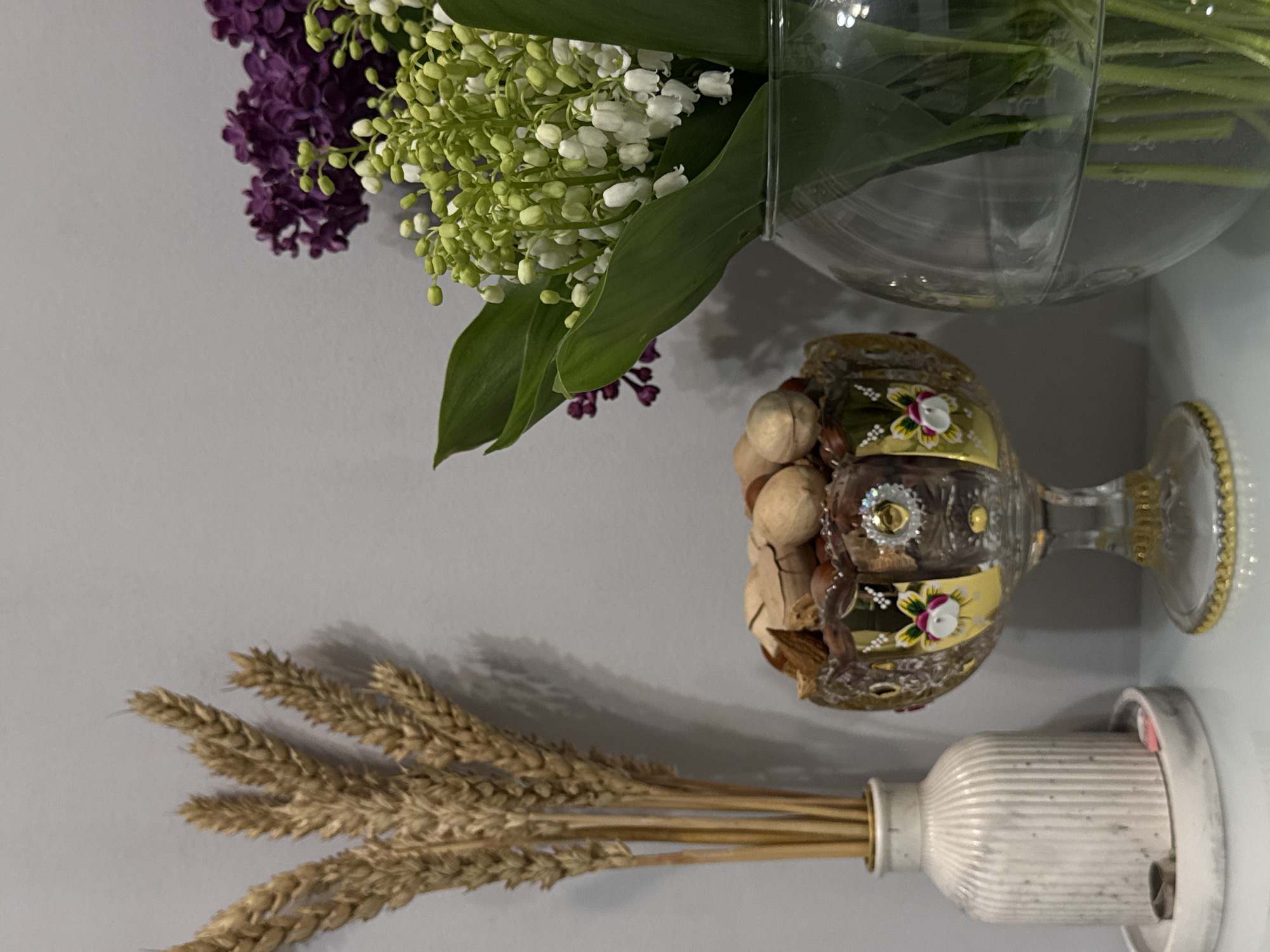 A still life composition showcases a collection of natural elements and decorative items arranged on a light surface against a plain wall. The image is presented with a 90-degree counter-clockwise rotation.
If oriented correctly, on the left, a bundle of dried, golden-brown wheat stalks is held in a white, fluted or ribbed ceramic vase. The individual ears of wheat are distinctly visible, casting soft shadows on the light grey background.
To the right of the wheat, a clear glass goblet with an ornate, gilded design is prominently featured. The goblet is adorned with gold patterns, painted pink and white floral motifs, and green leaves, resting on a gold-rimmed base. It is filled with a mixture of unshelled nuts, primarily walnuts, along with other dried botanical elements.
In the upper right section of the scene, a vibrant bouquet of fresh flowers is displayed in a clear glass vase. The bouquet consists of clusters of deep purple lilac blossoms and delicate white lily of the valley flowers, accompanied by their broad green leaves. The stems are visible through the transparent vase, which contains water.
The lighting is soft and even, suggesting an indoor setting. There are no people or visible text in the image.