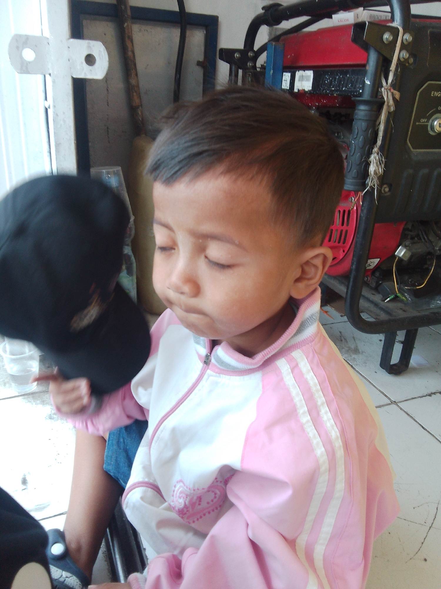 A young child, likely male, is seated in the foreground, facing partially away from the camera and looking down. The child has dark hair and is wearing a pink and white tracksuit with white stripes down the sleeves. The child's left hand is raised, holding a black baseball cap. Behind the child, a black generator with red accents is visible, along with a blue and white framed structure. The floor appears to be tiled. The background elements suggest an outdoor or semi-outdoor setting, possibly a workshop or storage area. The scene is set in the city of Cianjur, Indonesia.