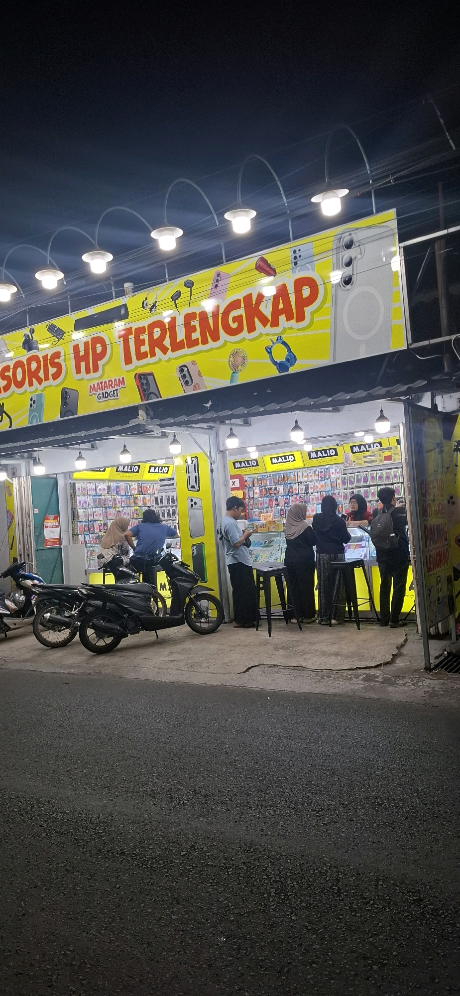 This is a nighttime, eye-level, vertical, outdoor shot of a storefront in Pabelan, Indonesia, advertising "Aksesoris HP Terlengkap" (Most Complete Phone Accessories). The shop is illuminated by bright yellow signage and multiple overhead lights. The main sign features images of various phone accessories, including headphones, cases, and a small fan. Below the main sign, the storefront itself is visible, with shelves displaying a wide array of phone cases. Several people are gathered inside and around the entrance of the shop. Two motorcycles are parked on the pavement in front of the store. The pavement itself appears cracked in places. The background shows a dark, unlit area, suggesting the shop is on a street in a built-up area.