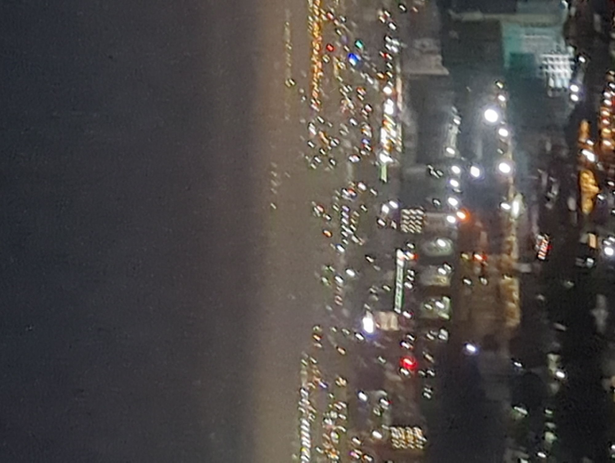 The image shows an aerial view of Bishkek City, Kyrgyzstan, at night. City lights illuminate buildings and roadways. To the left, a dark area possibly represents a park or undeveloped land. Streets are marked by the lights of moving vehicles. Tall buildings are identifiable by clusters of window lights.