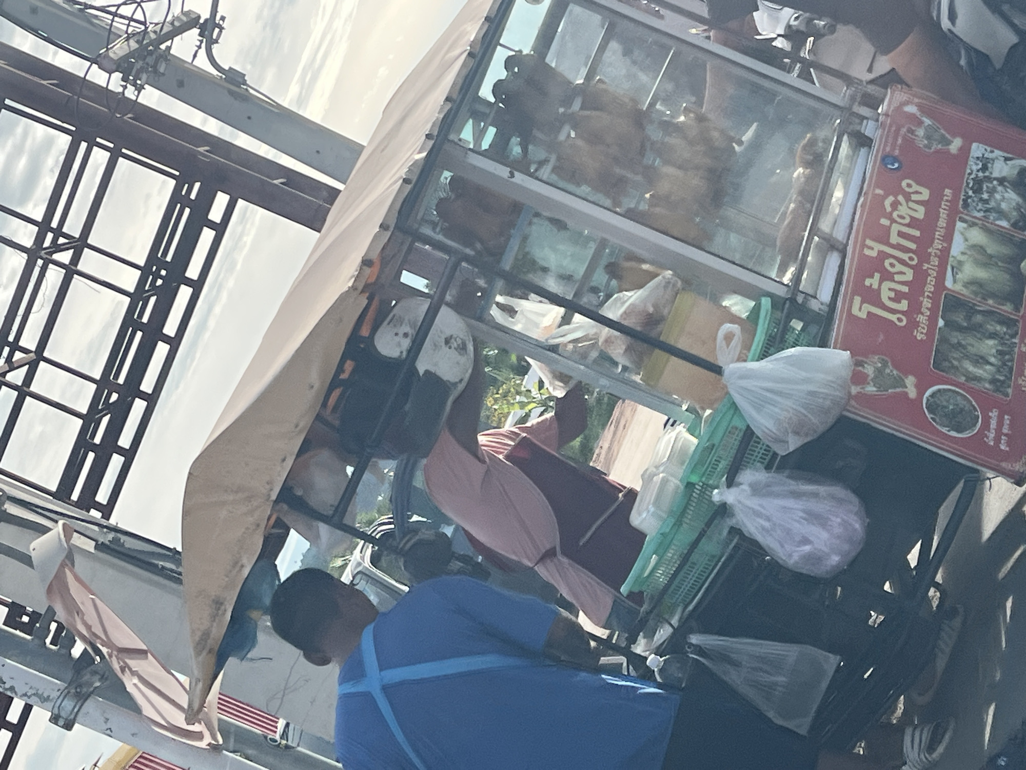 The image shows a street food vendor in Baan Bang Rak, Thailand. The vendor, wearing a blue shirt, is preparing food at a mobile cart with a glass display case, showcasing roasted chickens. The cart also has bags of food, possibly ready for customers. A faded awning provides shade. Power lines are visible above the cart.
