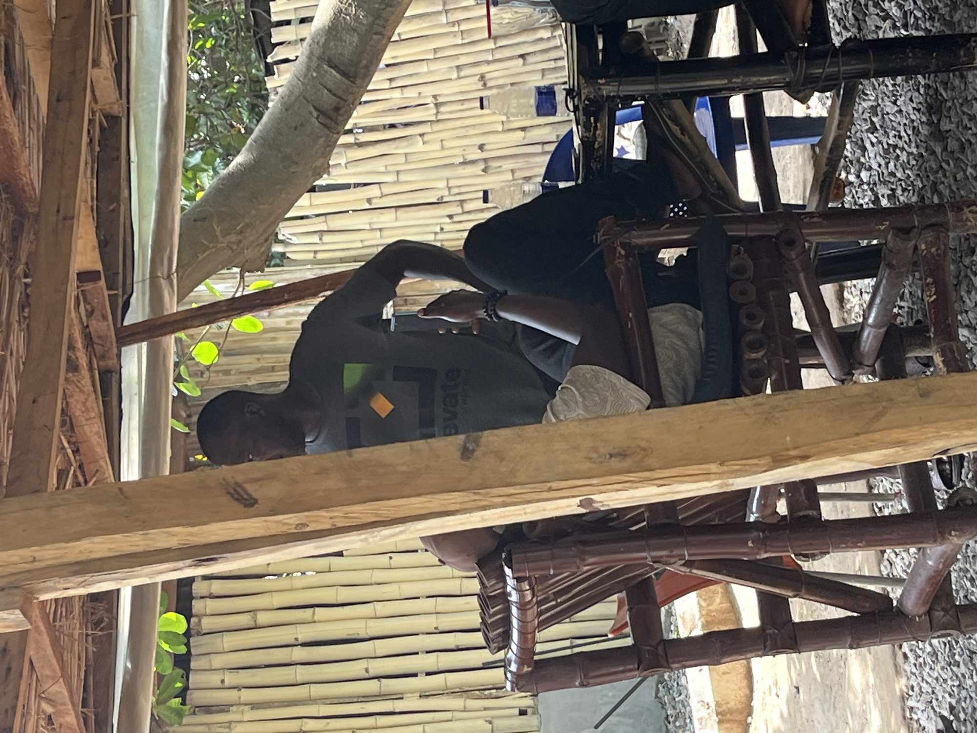The image shows a person lying down on a wooden structure, possibly a hammock or a raised bed, with their head resting on their arm. They are wearing a dark t-shirt with a graphic design and text on it. The background consists of bamboo walls and a tree branch. The scene appears to be outdoors, in a natural or rustic setting. The lighting suggests it could be daytime, with sunlight filtering through.
The text on the t-shirt is partially visible and reads "levate". The graphic design includes an orange square. The person's posture suggests relaxation or rest. The overall impression is one of a casual, tranquil moment in an outdoor environment. The location is stated to be Abuja, Nigeria.