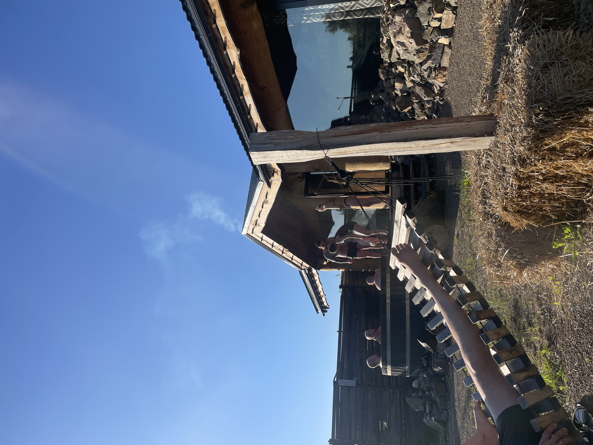The image, oriented correctly, displays an outdoor leisure scene under a bright, clear blue sky in Дрокино, Russia. The central focus is a rustic wooden building, possibly a sauna or bathhouse, with a prominent sloped roof, wooden beams, and a large reflective window that mirrors the sky and surrounding environment. Piles of rocks are visible near the building's foundation, and to the right, large bales of hay add to the rural aesthetic. The ground is a mix of gravel and sparse vegetation.
Several people are present, engaged in recreational activities. On a wooden deck or porch area connected to the building, three individuals are visible, appearing to be relaxing or socializing, dressed in swimwear or casual attire. To the lower right of the building, two more individuals are partially submerged in a large, dark wooden barrel structure, likely a hot tub or cold plunge pool, with only their heads visible above the rim. Wisps of smoke or steam rise from behind the main building, suggesting a sauna or heating process is underway.
In the immediate foreground, a pair of bare legs belonging to the viewer are stretched out on a slatted wooden lounger or bench, indicating a relaxed first-person perspective. The bright sunshine suggests it is daytime, casting long shadows and creating a sense of warmth and leisure. The clear sky and sunny weather contribute to a pleasant outdoor atmosphere. No visible text is present in the image.
