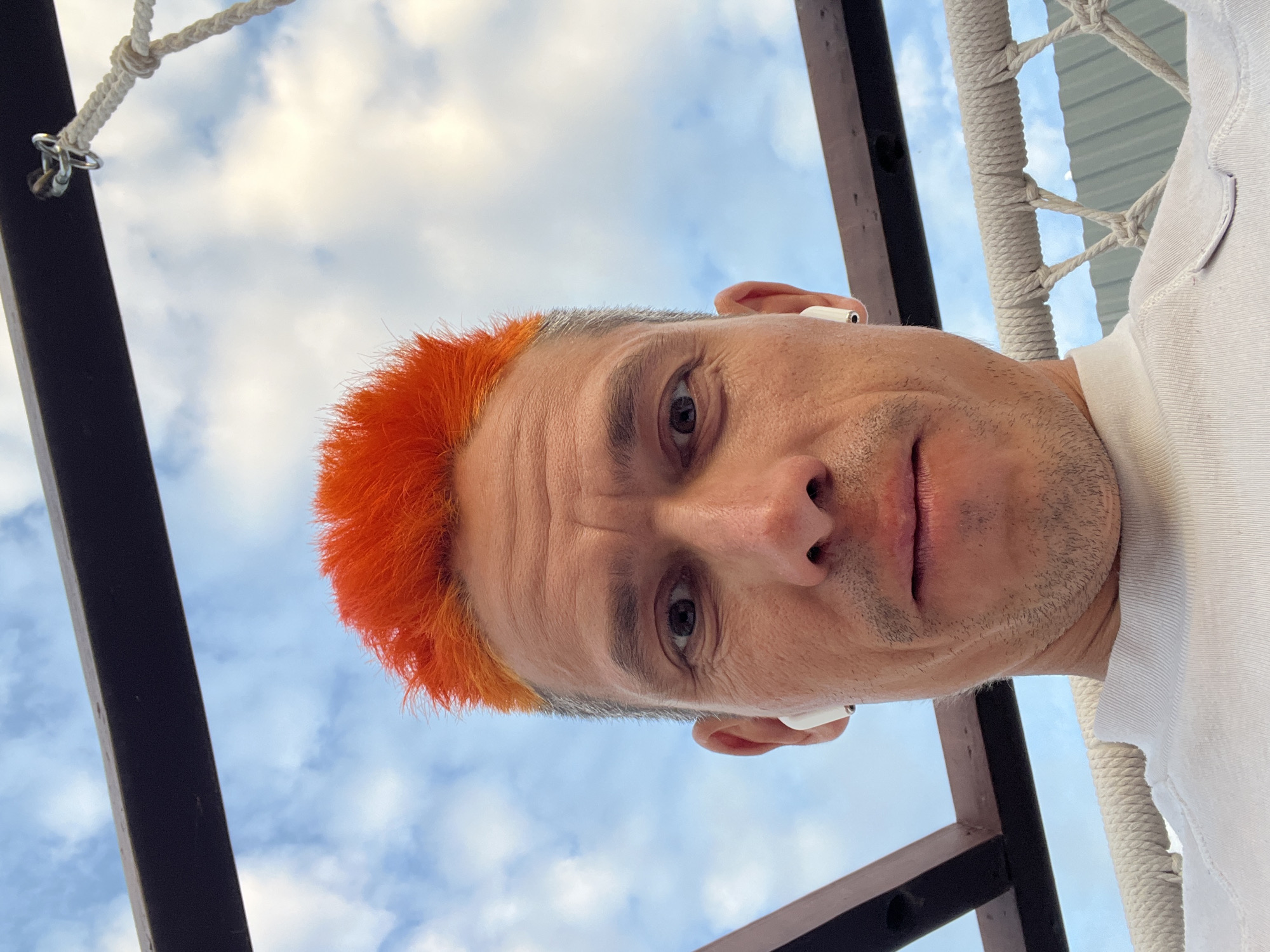 A low-angle, close-up shot of a man with a dyed orange mohawk, likely a selfie. He is wearing white earbuds. The background shows a partly cloudy sky and elements of an outdoor structure, potentially a swing or hammock, with a dark metal frame and rope supports. The photo appears to have been taken in an outdoor setting. The location, Chaikovsky, Russia is not discernible from the image.