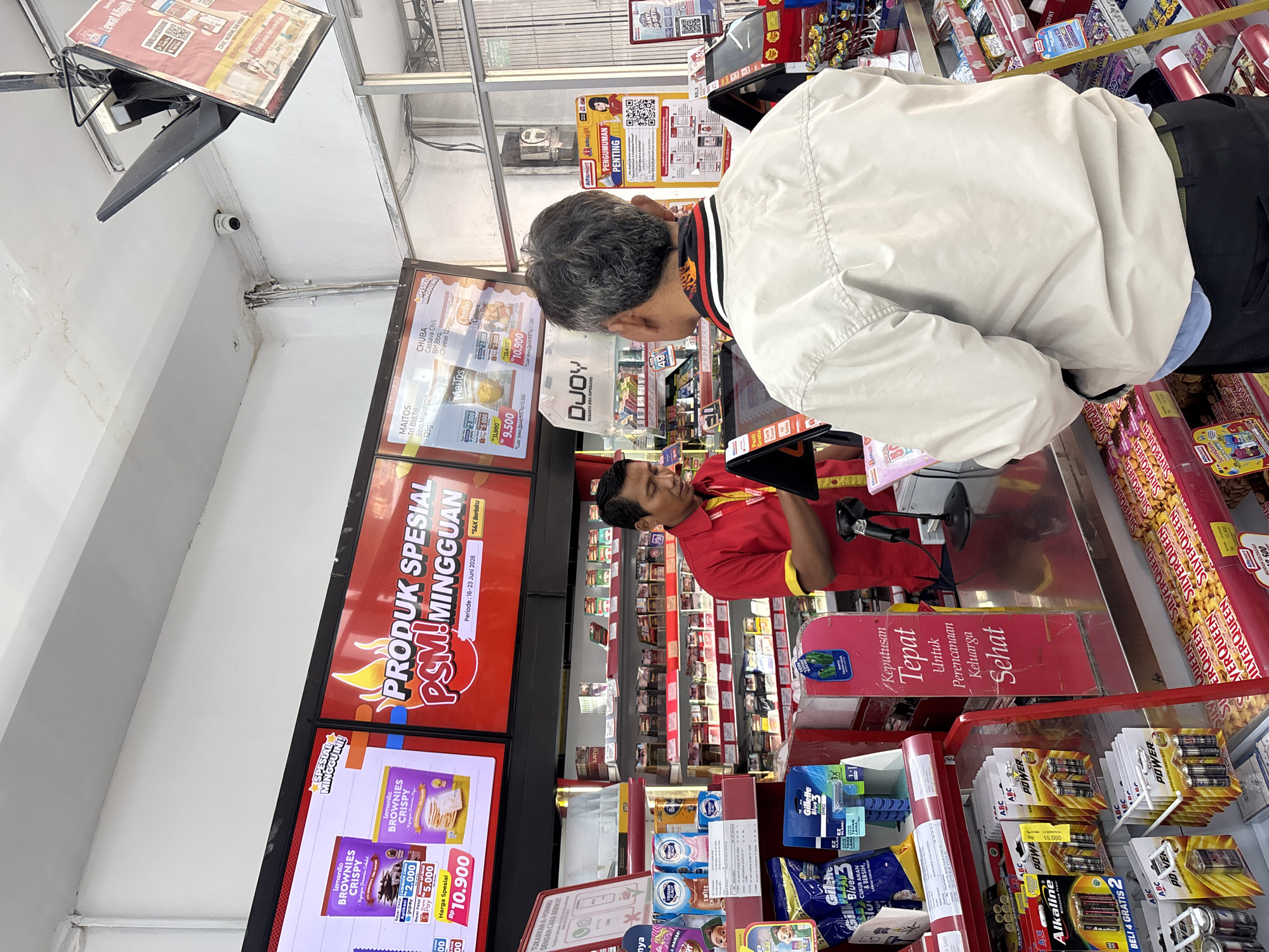 This is a medium shot, eye-level, indoor photograph taken in a convenience store, likely an Alfamart, in West Jakarta, Indonesia. The scene captures a transaction at the checkout counter.
In the foreground, a man with graying hair and wearing a light-colored jacket is standing with his back to the camera, facing the checkout counter. He appears to be engaged in a transaction.
Behind the counter, a male store employee in a red and yellow uniform is standing, looking down at a digital screen. He is in the process of serving the customer.
The checkout counter itself is cluttered with various items. To the left of the counter, there are shelves stocked with products such as batteries (ABC Alkaline Power), razors (Gillette), and other packaged goods. Behind the employee, shelves are filled with numerous cigarette packs.
Above the counter, digital screens display advertisements. One screen prominently shows "PRODUK SPESIAL PSM MINGGUAN" (Special Weekly Products) with fiery graphics. Another screen displays various product promotions, including "BROWNIES CRISPY" and snacks like "MAITOS" and "CHUBA".
To the right of the counter, a poster with the Alfamart logo is visible, featuring a QR code and information titled "PENTING" (Important). Another poster with the title "HANYA SO DETOX" is also present. The store environment appears well-lit with fluorescent lighting.
The overall impression is a typical retail setting in Indonesia where everyday consumer goods are sold.