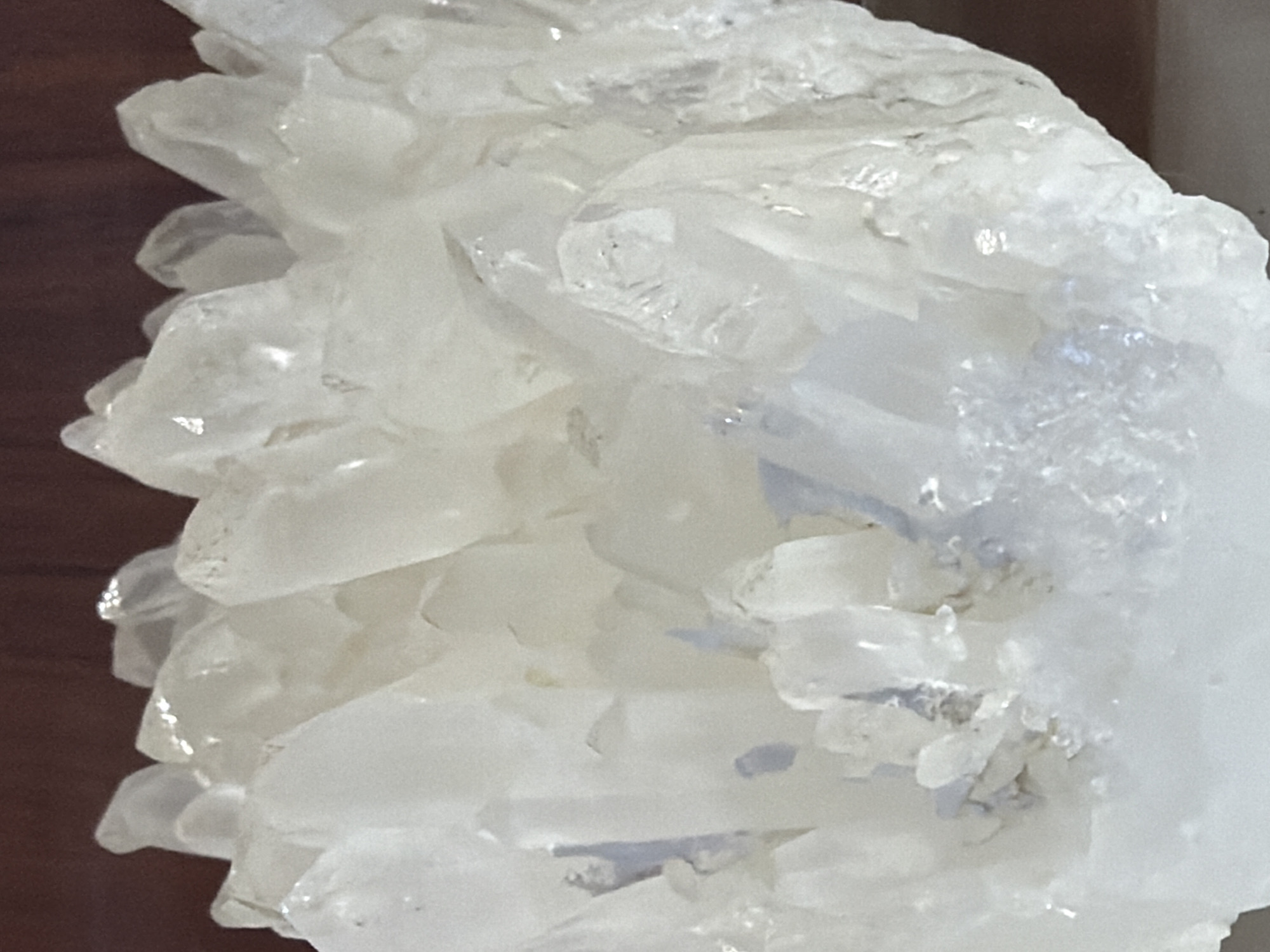 This is a close-up, medium-shot photograph of a cluster of clear, translucent crystals. The crystals are jagged and angular, with many facets reflecting light. They appear to be quartz or a similar mineral. The background is blurred and consists of a dark, grainy surface, possibly wood, on the left side, and a lighter, out-of-focus area on the right. There is no discernible activity or event taking place, as the image focuses solely on the mineral formation. The lighting is bright, highlighting the transparency and texture of the crystals. The image is devoid of any text or people. The location context provided is Akatovo, Russia, but there are no visual cues within the image itself to confirm this.