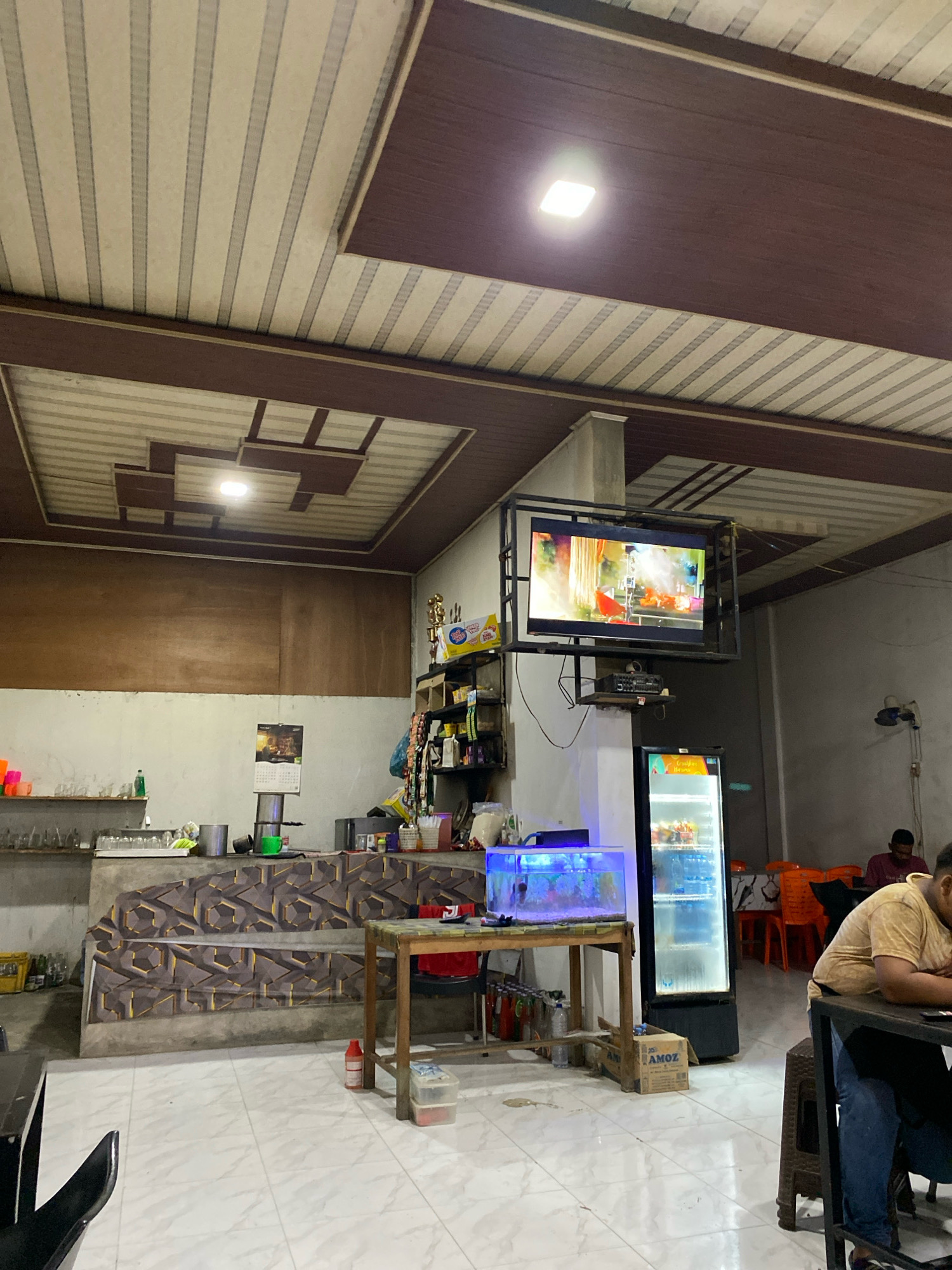 The image shows the interior of a restaurant or cafe. In the foreground, a person wearing a yellow shirt and blue jeans is seated at a table, looking down at their phone. To the left of this person is a black chair. Further into the scene, there is a wooden table with a glass-top aquarium containing fish and blue lighting. Next to the aquarium is a tall, narrow refrigerator filled with beverages, with a television mounted on the wall above it. In the background, there are tables and chairs, and a person is visible sitting at one of the tables. The ceiling is paneled with a brown and beige design, and there is a square light fixture. The walls are a light color, with a wooden paneling on the upper section. A calendar is hanging on the wall. The floor is tiled with a white marble pattern. The overall impression is that of a casual dining establishment. The image is consistent with a location in Lhokseumawe, Indonesia, given its common architectural and interior design features for such establishments in the region.