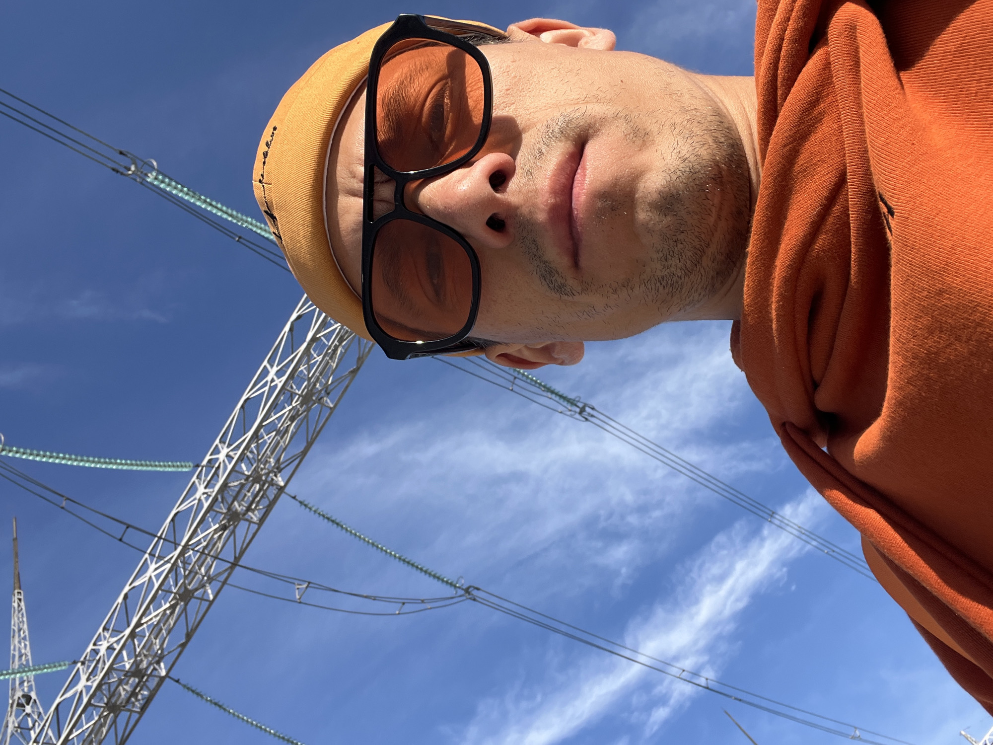 A low-angle, inverted close-up captures a person looking directly downwards towards the camera, set against a vibrant blue sky dotted with soft, wispy white clouds. The subject wears dark-framed sunglasses with distinct orange-tinted lenses, an orange beanie, and an orange hooded sweatshirt. Their face shows light stubble, and their lips are slightly parted, conveying a neutral expression. The unusual perspective places the viewer beneath the person, creating an intimate yet detached gaze.
In the background, prominent elements of an industrial landscape are visible: a large metal electricity pylon (transmission tower) and multiple power lines with characteristic green/transparent glass or ceramic insulators. This outdoor scene, bathed in bright daylight, is situated in Chaikovsky, Russia. Faint, dark cursive text is visible on the side of the orange beanie, though it is illegible. The image appears to be a direct selfie or portrait taken from a unique, upward-looking vantage point.