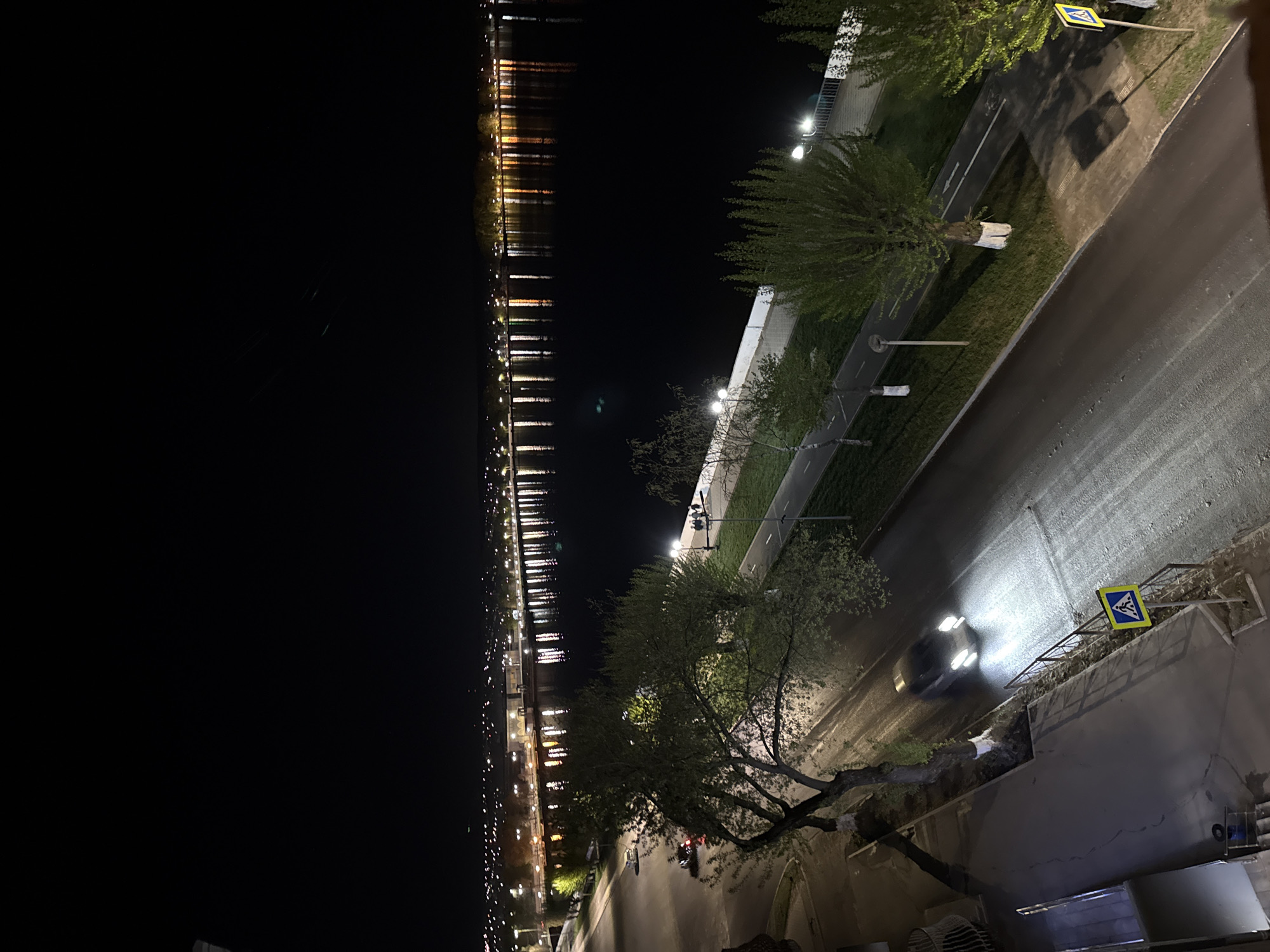 The image depicts a nighttime scene in Votkinsk, Russia, likely taken from an elevated perspective such as a high-rise building. The view encompasses the city's waterfront, identifiable by the string of lights reflecting on the surface of the Votkinsk Reservoir. A road runs alongside the reservoir, with trees and vegetation lining the walkway. A pedestrian crossing sign is visible next to the road. A car is driving along the road, its headlights illuminating the pavement. The time of day is night, as indicated by the darkness and artificial lighting.