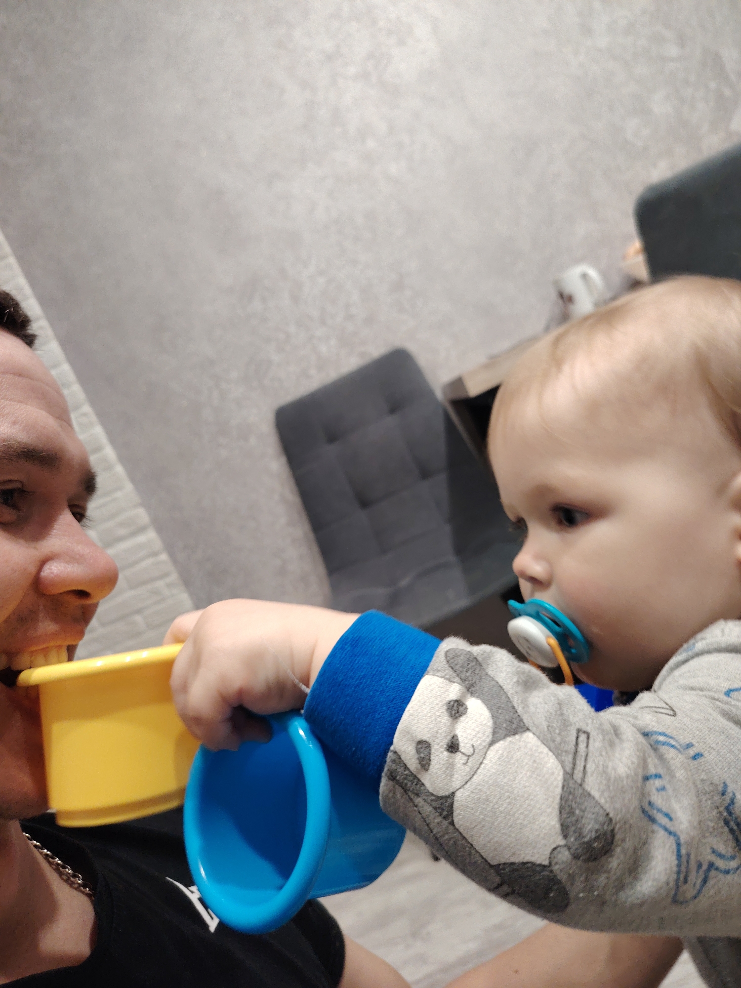 The image depicts an adult male with short brown hair, wearing a black t-shirt and a silver chain, holding a yellow cup in his mouth. A toddler with a pacifier in their mouth is offering a blue cup towards the adult's face. The child is wearing a gray sweatshirt with a panda design and blue cuffs. The background includes a gray wall, a gray chair, and a small table with a white mug on it. The image was taken indoors in what appears to be a home setting. The location is not identifiable in the picture, it is unknown if it was taken in Chaikovsky, Russia.