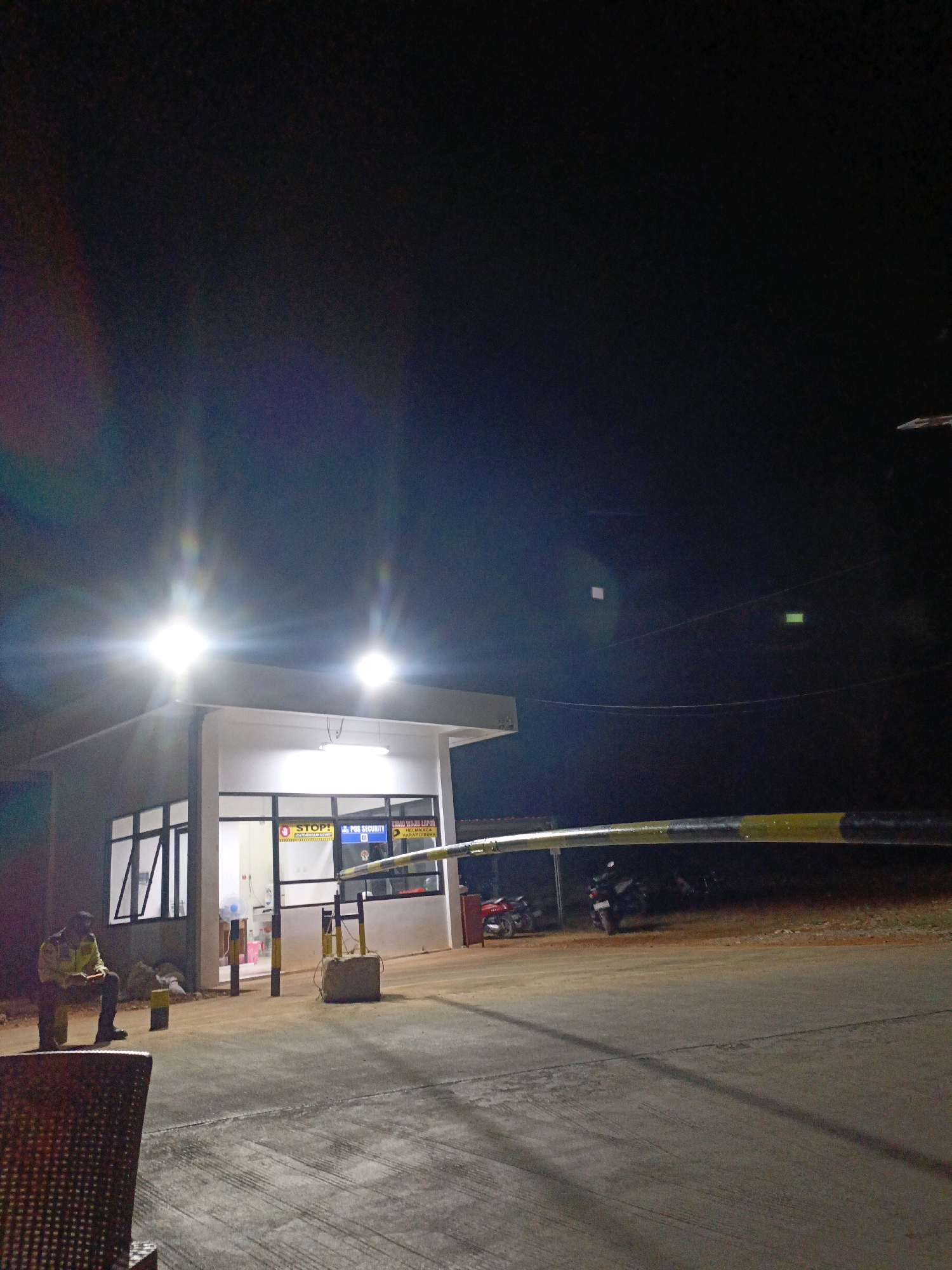 The image depicts a security post at night, likely in Jenu, Indonesia. A single security guard, wearing a yellow uniform, is seated on a bench outside the post. The post is a small, white building with large windows. A yellow and black striped barrier is raised, indicating it is open. Several motorcycles are parked in the background, some of which are illuminated by the post's lights. The scene is lit by bright, artificial lights that create lens flares. The ground is paved and appears to be a road or parking area. There are electrical wires visible in the dark sky above.