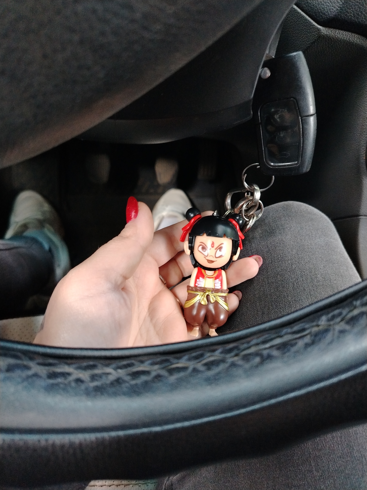 A woman is holding a keychain with a doll figure inside a car. The keychain is attached to the car key. The woman's hand with painted nails holds the doll keychain. The background includes the car's dashboard, steering wheel, pedals, and the woman's lap with gray pants. There is no information about the location of Yaroslavl, Russia.