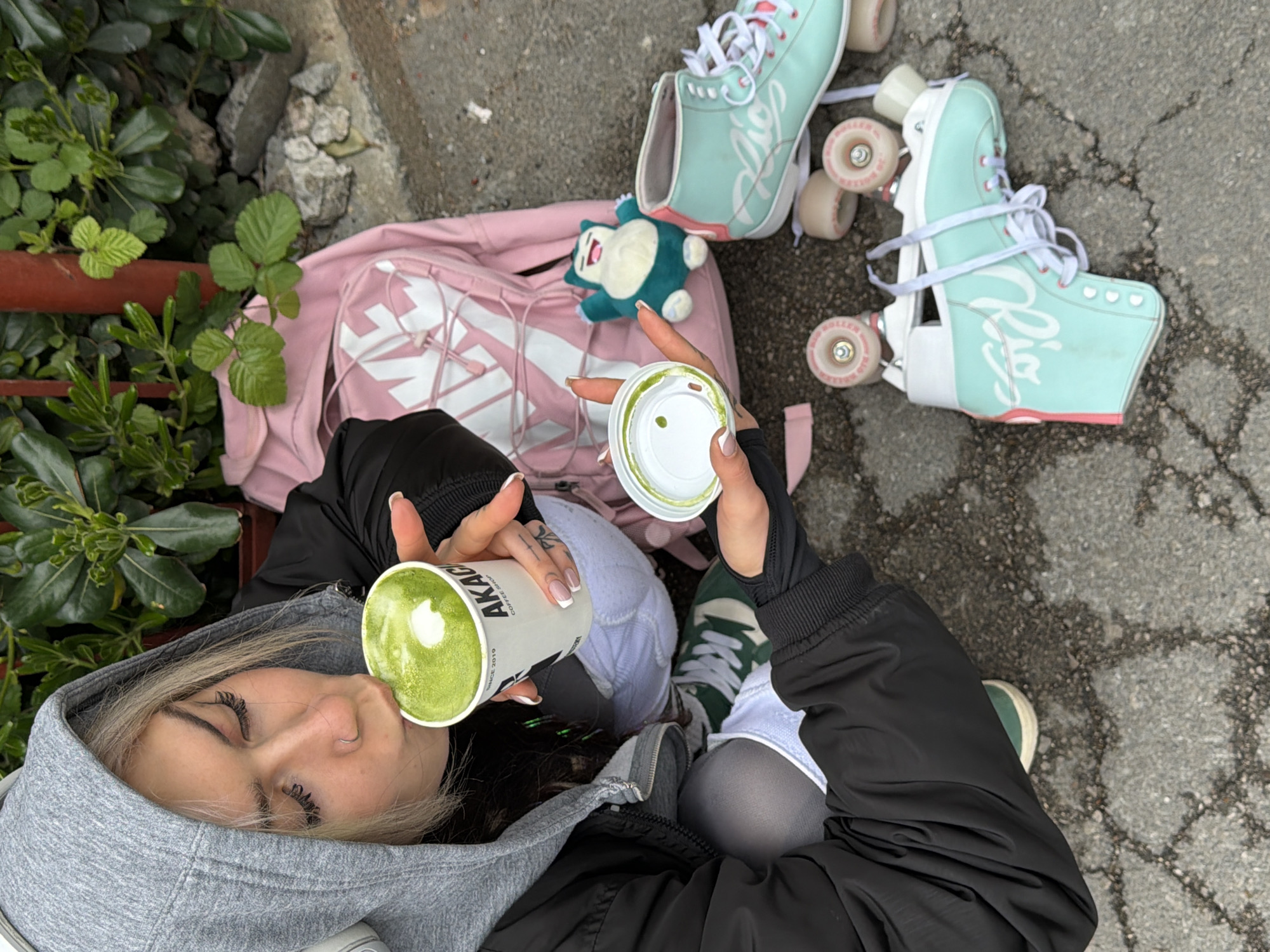An individual, identified as a young adult female, is depicted seated on a grey asphalt surface in Budva, Montenegro. She wears a grey hooded sweatshirt with the hood up, a black zip-up jacket, light-colored shorts over dark grey tights, and green and white sneakers. Her blonde hair is visible, and she has a nose piercing (stud) and French-tipped manicured fingernails. Tattoos are present on her left hand/wrist.
The individual is actively drinking from a white paper cup, branded "AKACIA COFFEE SHOP," which contains a green, foamy beverage with a distinct white circle on its surface, consistent with a matcha latte. Her eyes are directed downwards towards the cup. In her right hand, she holds a white plastic lid, which shows green residue on its interior surface.
To her left and slightly behind her, a pink backpack with a prominent white logo is resting against a dense green hedge. Next to the backpack, a small blue and white Snorlax plush toy is situated. A pair of teal and pink "Rio Roller" brand roller skates, equipped with white laces and pink wheels, lie on the asphalt to the right of the individual. White headphones are visible around her neck, beneath the hoodie.
The immediate environment consists of a cracked grey asphalt or concrete ground surface. In the background, there is a dense green leafy hedge, with a reddish-brown vertical pipe or post visible within it. Scattered pieces of broken concrete are present at the base of the hedge.