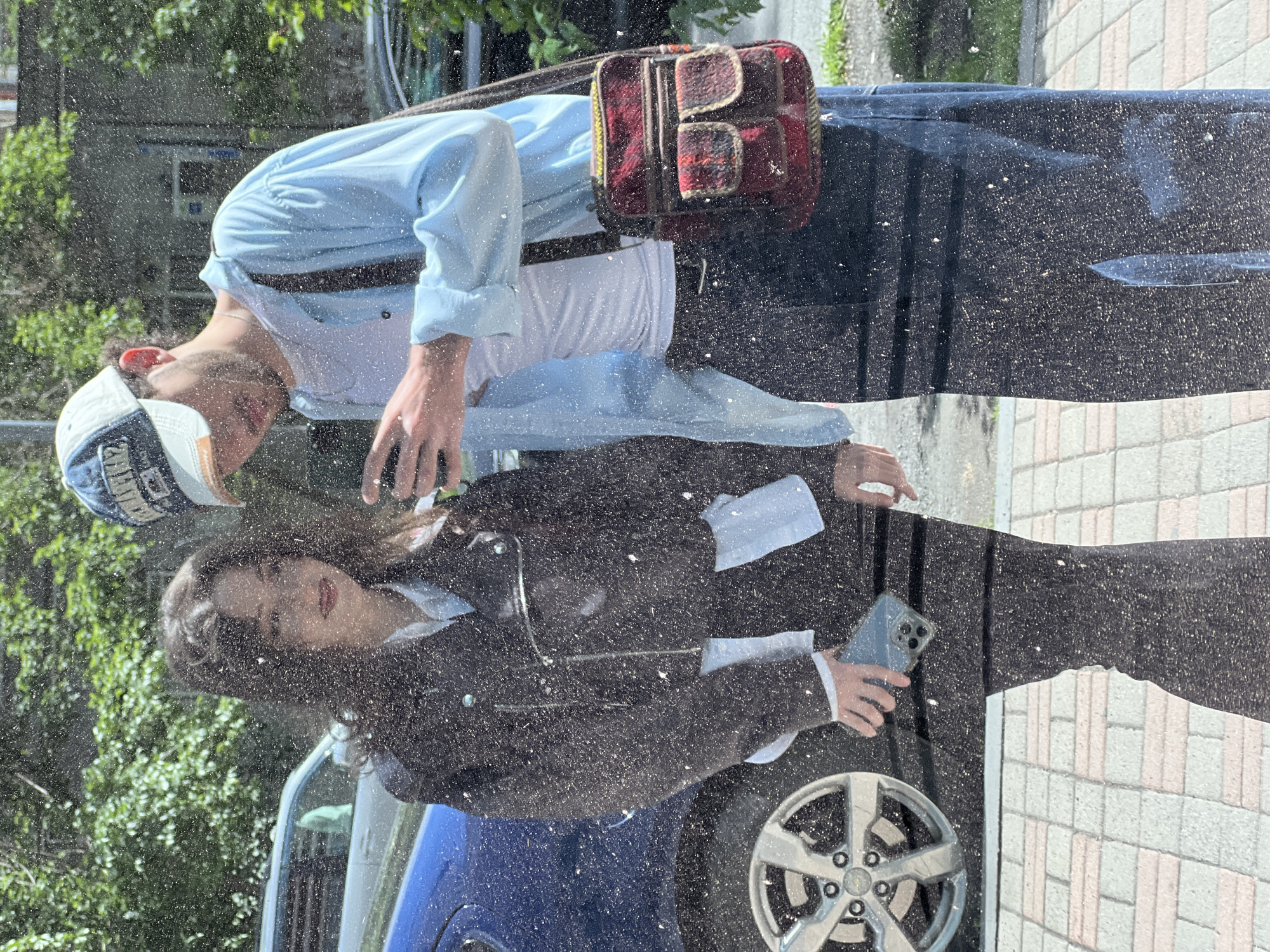 The image shows a reflection in a car window, obscured by water droplets. In the reflection, two people are visible. A man is standing on the left, wearing a light blue shirt, white t-shirt, dark jeans, a baseball cap, and a shoulder bag. A woman is standing on the right, wearing a black blazer over a white shirt and black trousers. She is holding a phone. The car is parked on a street with a brick-paved sidewalk. A blue car door and silver wheel are visible. Green trees are visible in the background, possibly reflecting the city of Yerevan, Armenia.