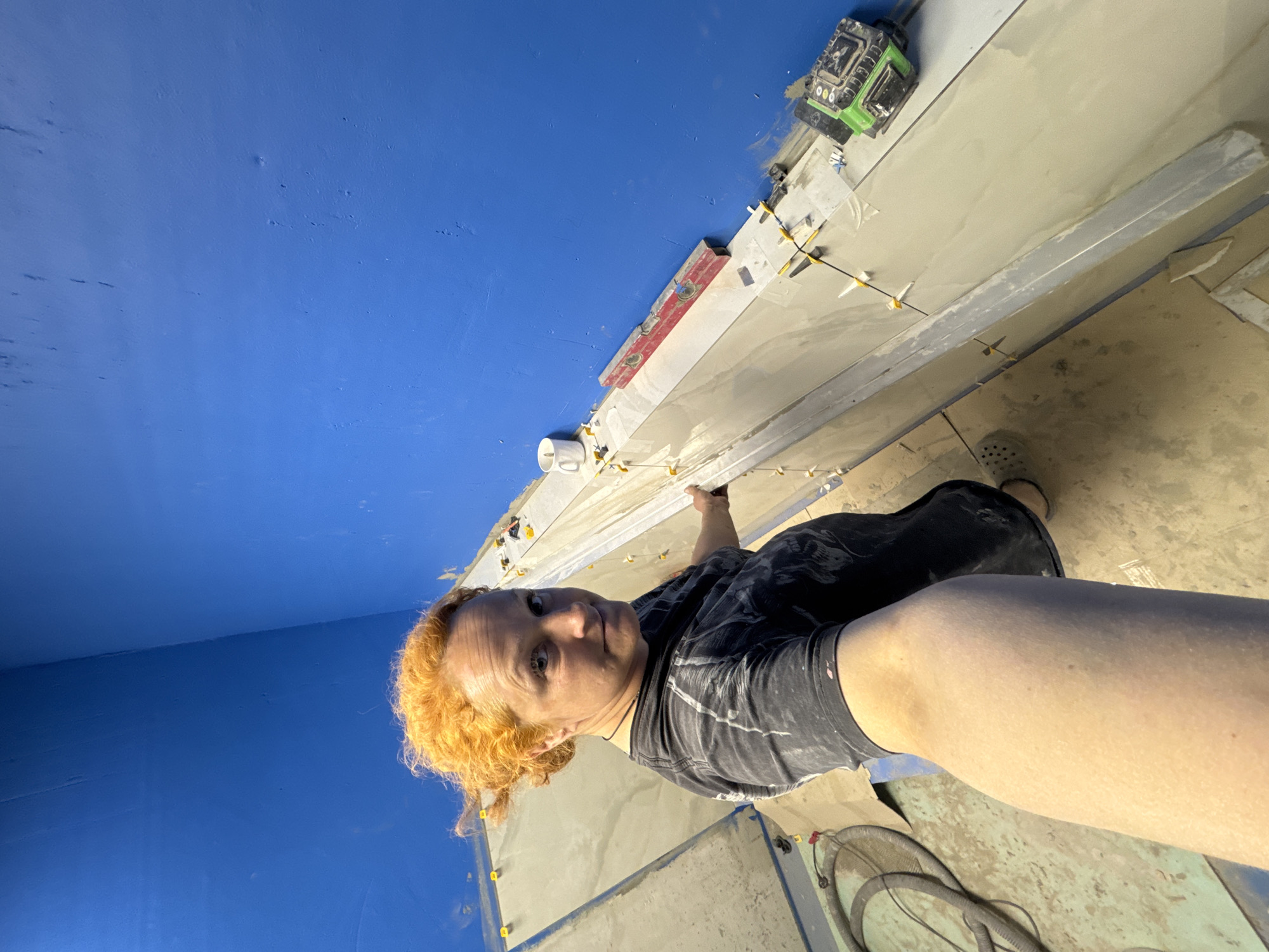 A waist-up selfie depicts a person with red hair, wearing a dark t-shirt, in a room with a bright blue wall on the left and tiles being installed along the top portion of the frame. The person's face is directed towards the camera. White tiles are visible, separated by yellow tile spacers. A laser level and another unidentified tool are situated on the tiled surface. A section of the floor is visible in the lower right corner, where a vacuum cleaner can be partially seen. The location is in Chaikovsky, Russia.