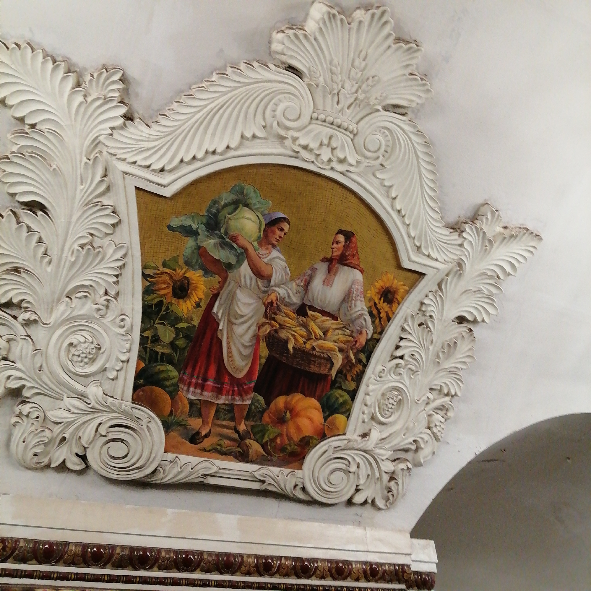 This image shows a mosaic panel framed with intricate white plasterwork. The panel depicts two women in traditional Ukrainian clothing in a rural setting. One woman carries a large cabbage, while the other holds a basket full of corn. Sunflowers, pumpkins, and watermelons are also present. The mosaic is set against a background of golden-colored tiles. The plasterwork surrounding the panel features stylized leaves, scrolls, and other decorative elements. The setting is most likely the Kievskaya station in the Moscow Metro, Russia.