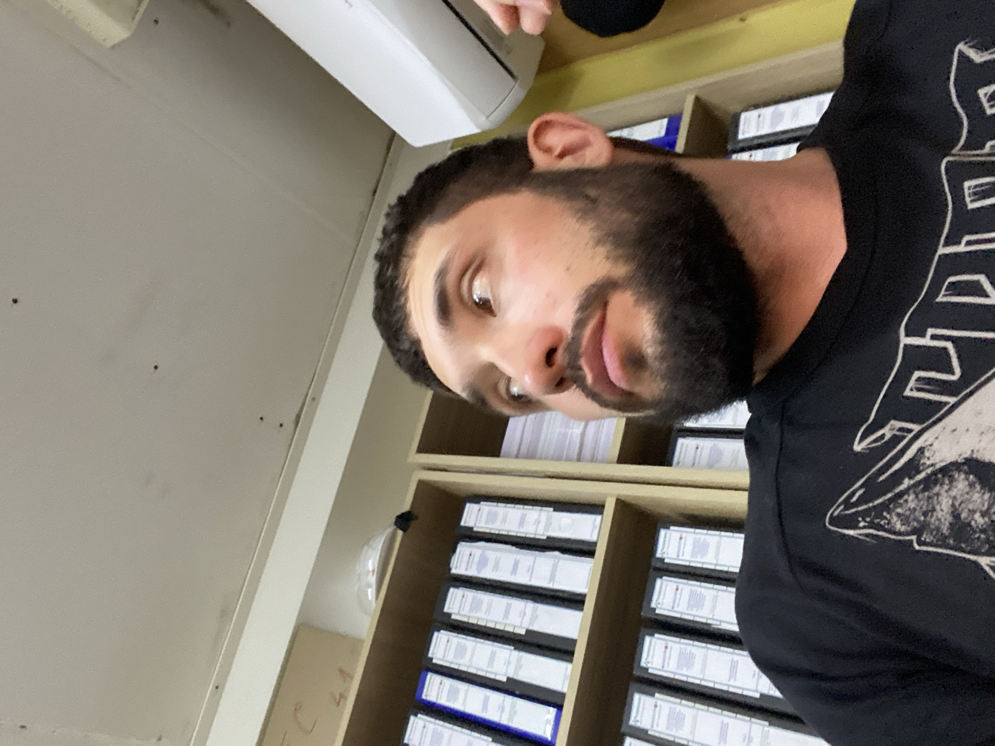 The image shows a close-up, low-angle, head-and-shoulders shot of a young adult male with dark hair, a beard, and brown eyes, looking to his right. He is wearing a black t-shirt with a graphic design. The background is an office environment with shelves filled with binders and folders. An air conditioning unit is visible on the wall above and to the right of the man's head. The lighting suggests an indoor setting with overhead lights. The image does not provide any information that would allow for the determination of the specific location within the city of Luzhitsy, Russia.