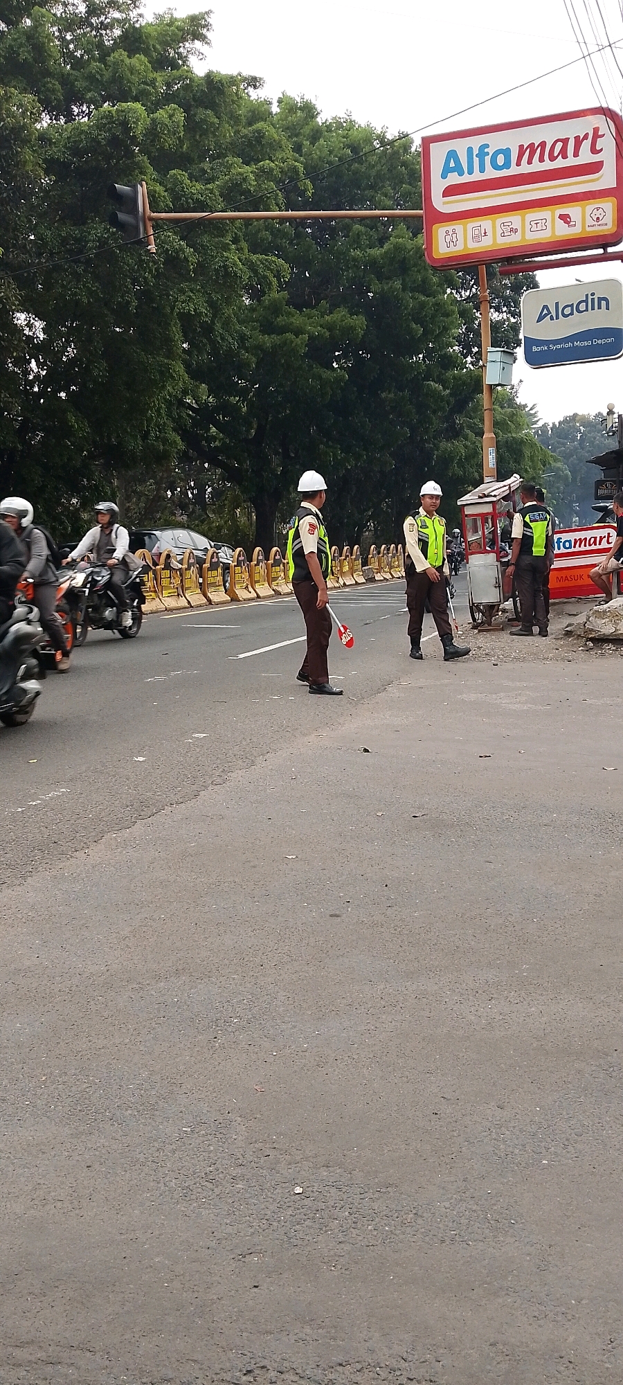 The image captures a street scene in Depok, Indonesia, near an Alfamart convenience store. The foreground features a paved road with some debris. In the middle ground, several individuals are present. Two traffic officers in light brown uniforms and reflective vests stand on the road, one holding a red "STOP" sign. They appear to be directing traffic, although no vehicles are directly interacting with them at this moment. Behind them, a row of yellow traffic barriers lines the side of the road. To the right, a small food cart is parked next to a pole. A sign for "Aladin Bank Syariah Masa Depan" is visible above the cart. Further back, motorcycles are seen on the road, with riders wearing helmets. In the background, a dense canopy of green trees dominates the scene, with a traffic light visible on a pole extending over the road. The sky is overcast.