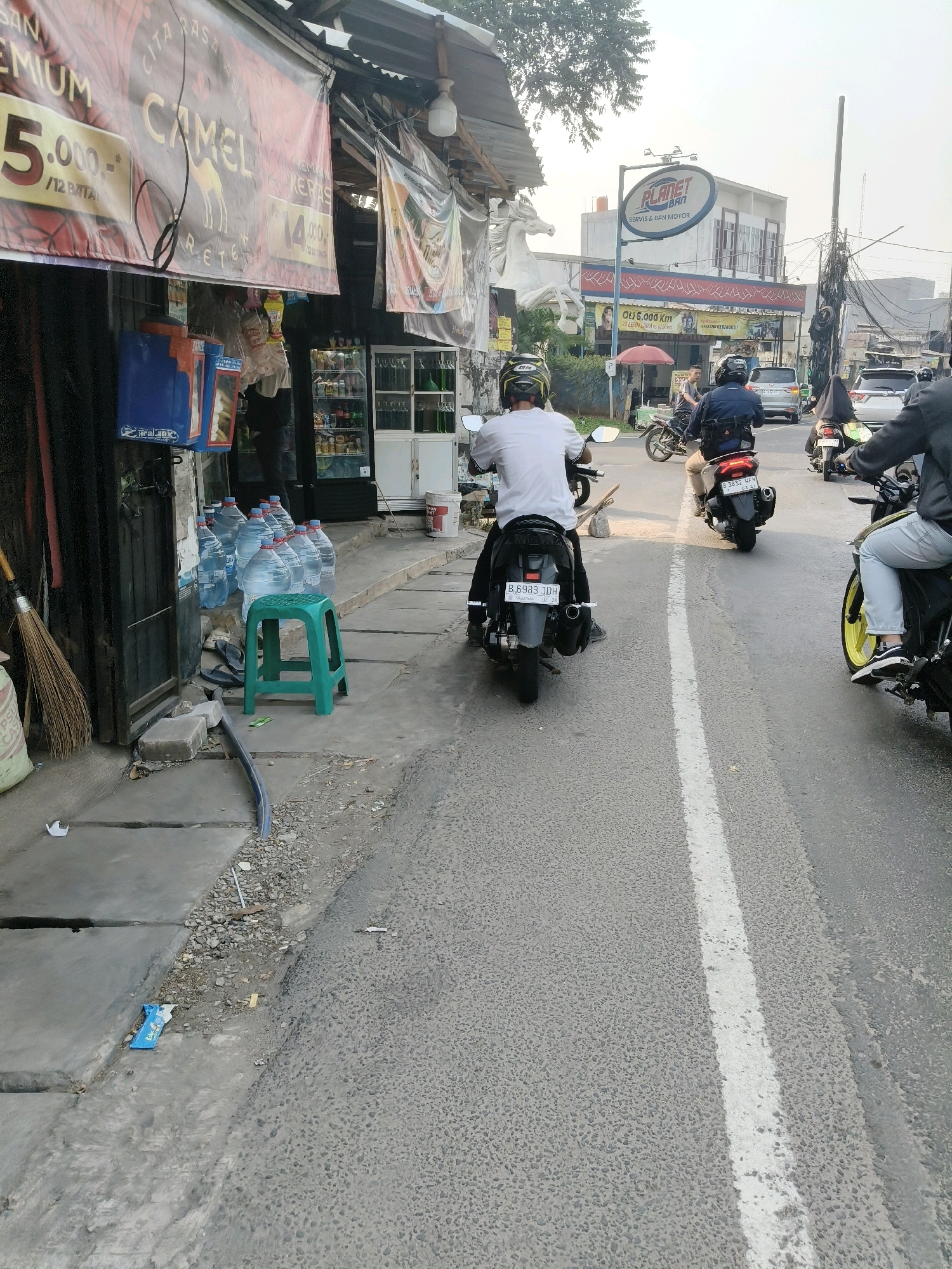 The image captures a street scene in South Tangerang, Indonesia, likely on a sunny day. In the foreground, a person on a motorcycle wearing a white t-shirt and a helmet rides away from the camera. To the left, a sidewalk area is visible with a green plastic stool, several large bottles of water, and a broom leaning against a dark wooden structure. Behind the bottles, a cooler box is visible. Further down the street, traffic is moving, with several other motorcycles and a car on the road. A sign for "PLANET BAN" (a motorcycle repair shop) is visible in the background, along with buildings and overhead power lines. The overall impression is that of a busy urban environment.