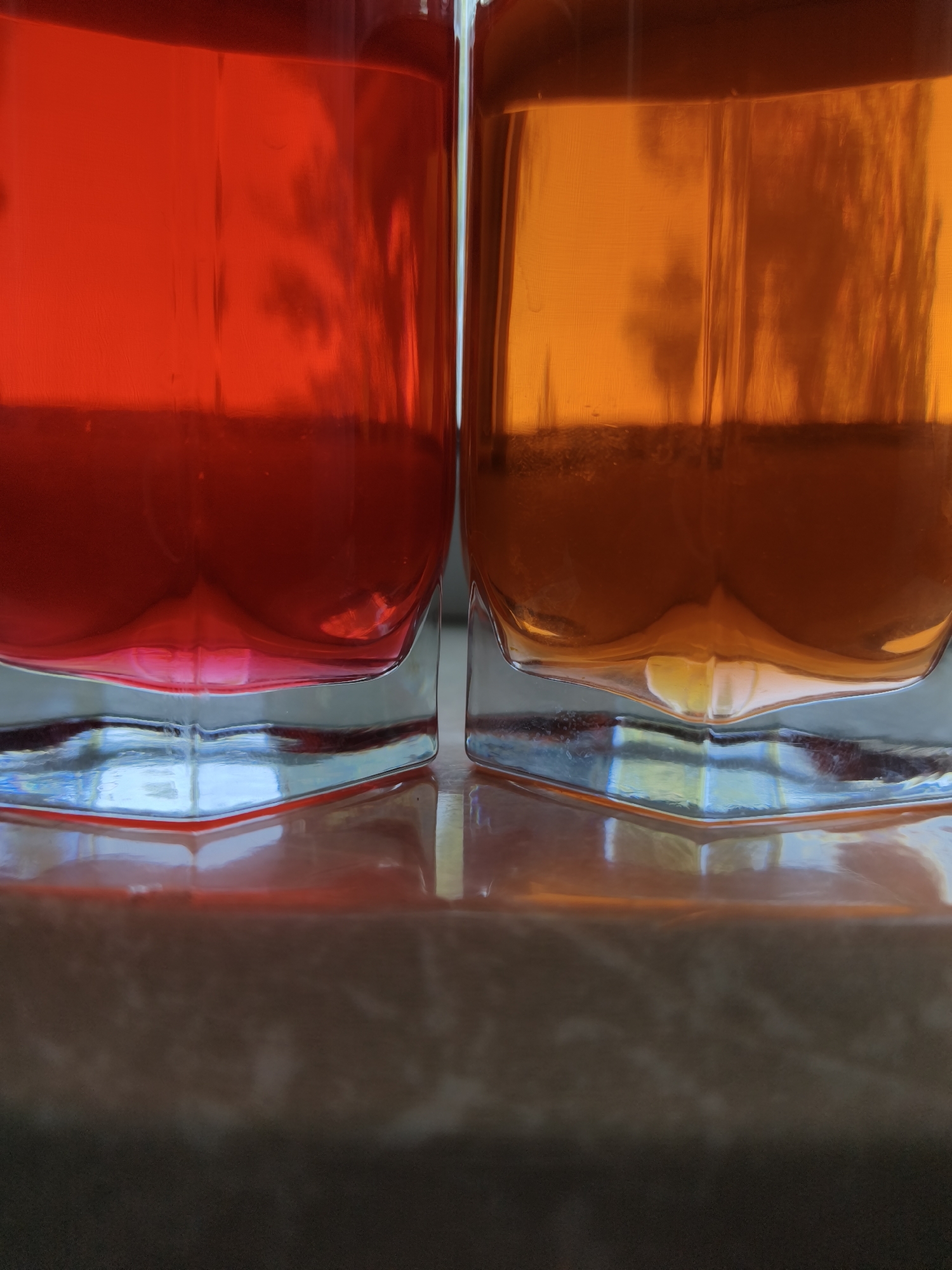 Two square glasses, one filled with a red liquid and the other with an orange liquid, are placed side-by-side on a reflective surface. The glasses are clear with geometric designs at the base, reflecting light and color. The surface below appears to be a mottled gray or brown, possibly a countertop or table. No people are visible in the frame. There is no explicit information to confirm the location in the city of Kryvyi Rih, Ukraine.