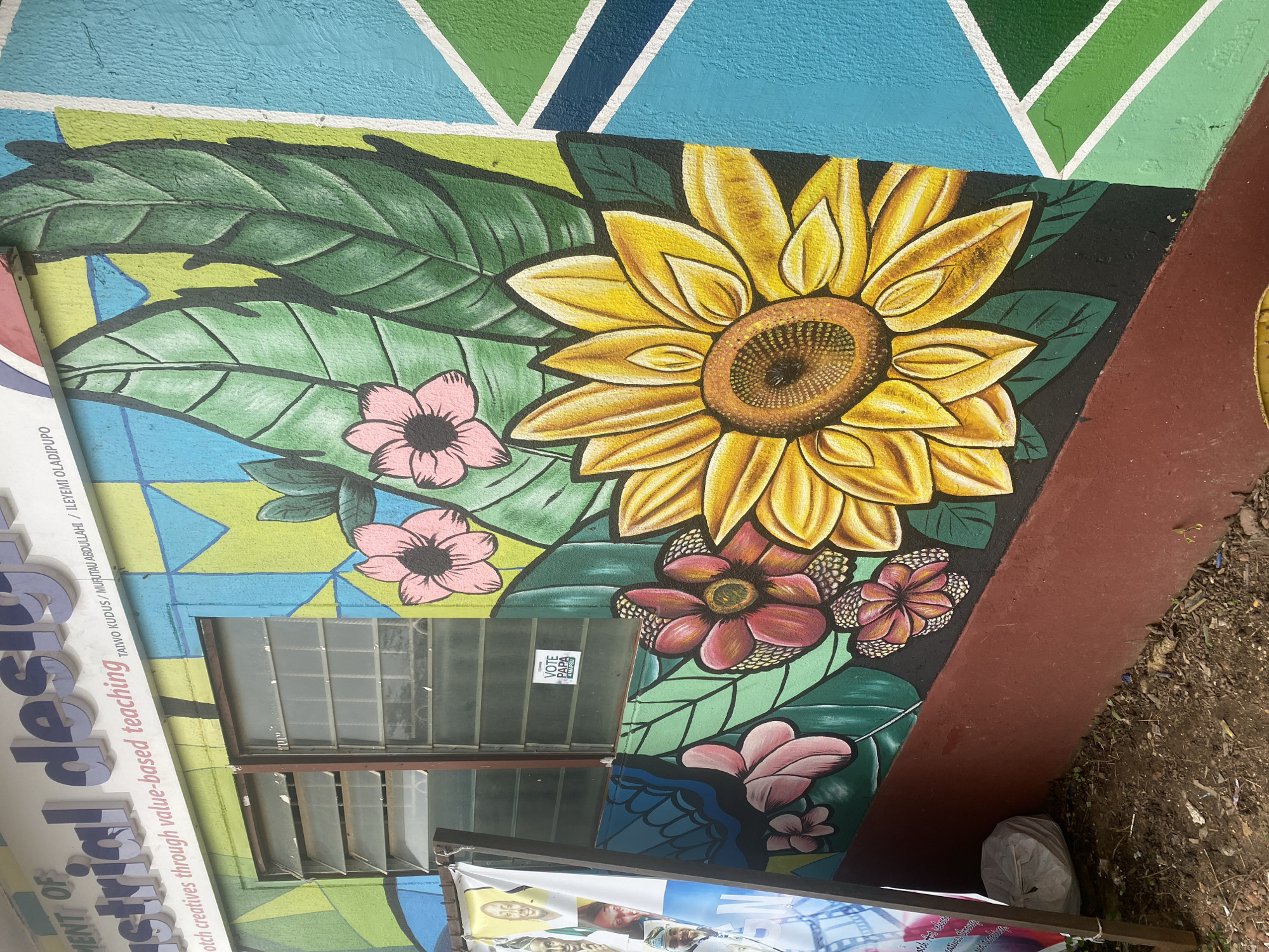 The image depicts the exterior wall of a building adorned with a vibrant mural in Asha, Nigeria. The mural features large, stylized flowers, including a prominent sunflower and several pink and red blossoms, interspersed with green leaves. The background is a dynamic geometric pattern of blue, green, and yellow. Above the mural, lettering on the building's facade reads "DEPARTMENT OF Industrial design" and "tch creatives through value-based teaching." A small sticker on a windowpane displays "VOTE PAPA 4 MARCH." To the left, a banner with portraits of men is partially visible, attached to a wooden support. The ground in front of the building is dirt with scattered debris.