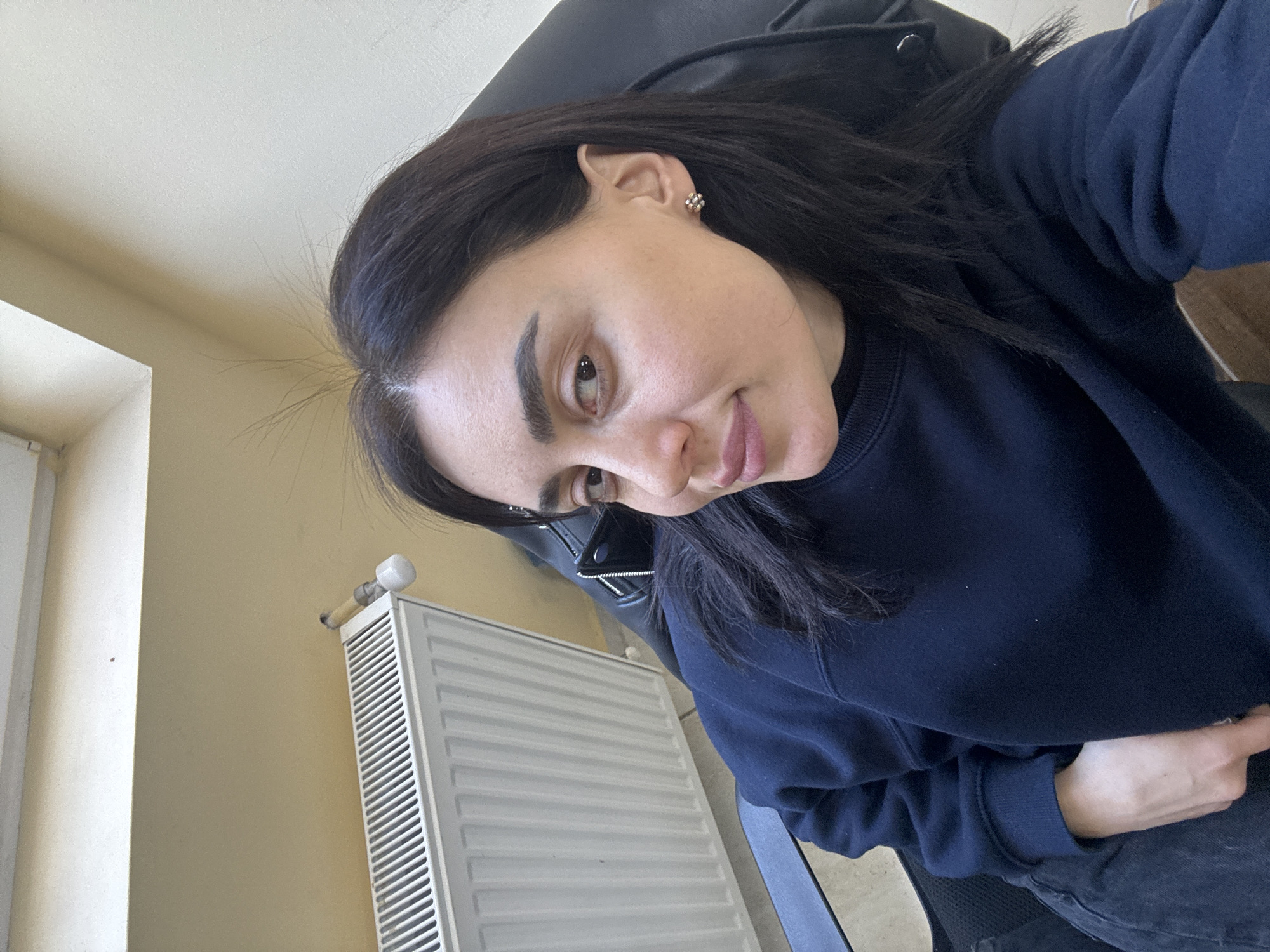 A woman with dark hair and eyebrows, wearing a dark blue sweatshirt, is pictured in an indoor setting. She is seated in a black office chair. Beside her is a white radiator. The wall is painted a light yellow. The location is assumed to be in Tbilisi, Georgia, although there is no specific visual evidence within the image to confirm the location.