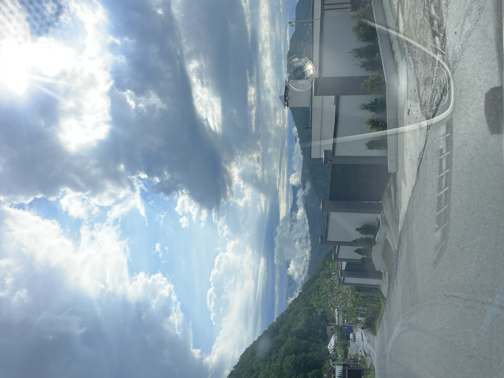 The image shows a view from what appears to be a vehicle in the city of Sochi, Russia. The sky is predominantly cloudy with patches of blue sky and bright sunlight. Distant mountains covered in green vegetation are visible. A modern building with a grey and white exterior runs along the right side of the image, reflected in the vehicle's window, along with what seems to be a street or road. In the lower portion of the image, the green hillside slopes down towards what might be residential or commercial structures.