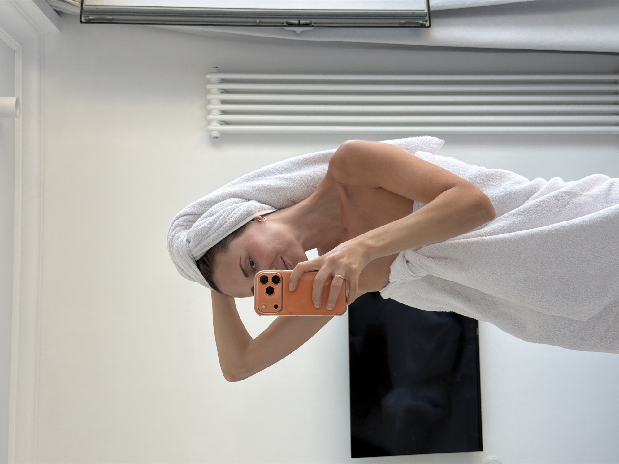 A woman is taking a mirror selfie in what appears to be a bedroom. She is wrapped in a white towel and has a towel on her head. She is holding an orange smartphone in her right hand. The background includes a black television, a white radiator, and white curtains. There is no visible indication of the city of Saratov, Russia.