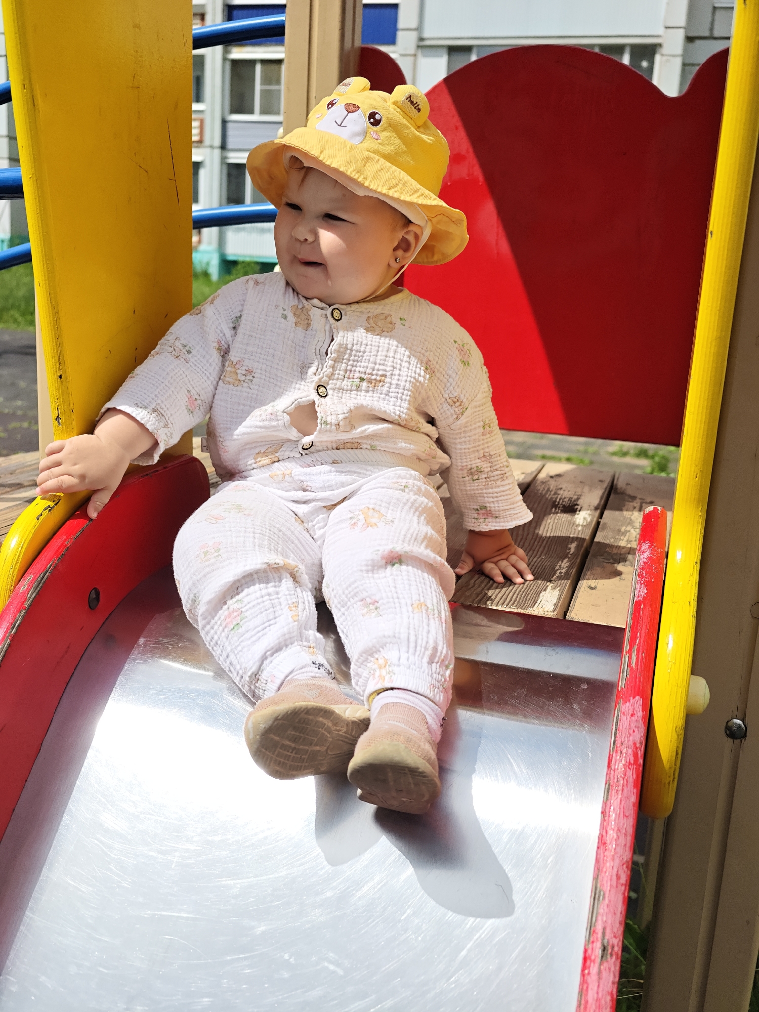 A toddler, wearing a yellow bucket hat with animal ears and a white patterned outfit, sits on a metal slide at a playground. The slide is part of a yellow and red playground structure. The background shows apartment buildings and greenery. The scene appears to be in the city of Chaikovsky, Russia, likely during daytime and fair weather.
