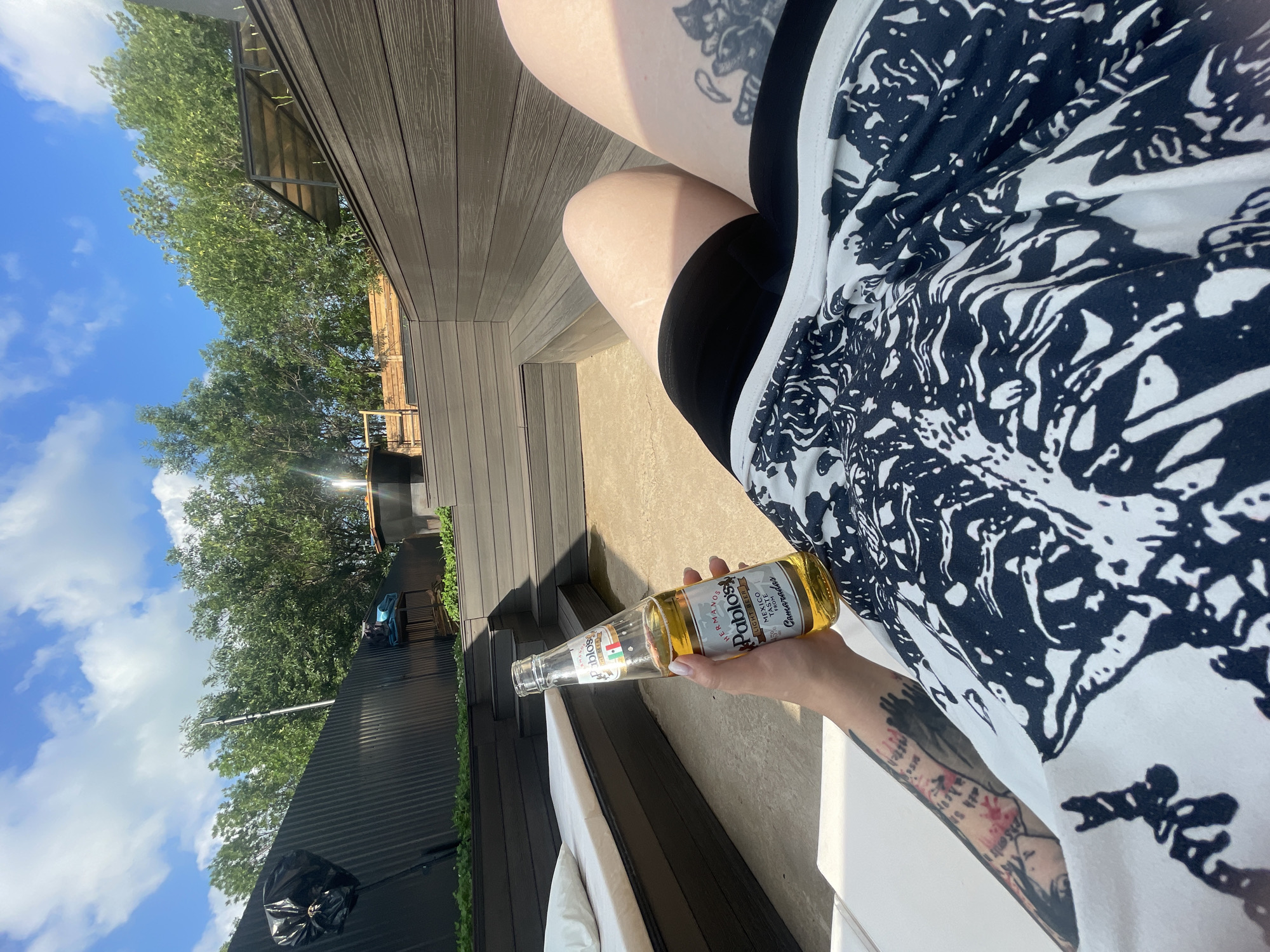 A person is relaxing outdoors, holding a bottle of beer and wearing black shorts and a black and white patterned shirt. They are sitting on a concrete patio next to a lounge chair with a white cushion. In the background, there is a dark wooden deck with built-in seating, a black corrugated metal fence, and trees under a blue sky with white clouds.