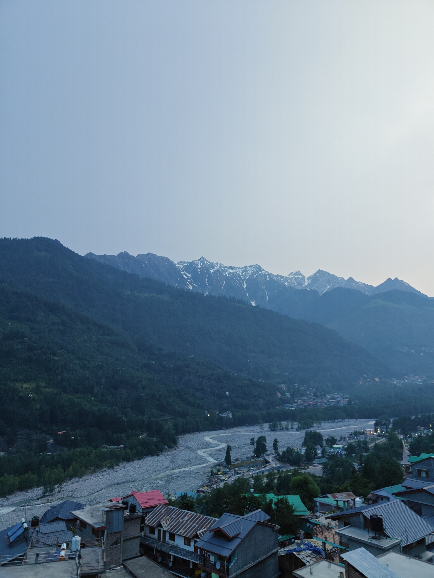 This is an eye-level, wide shot photograph of the city of Vashisht, India, taken from a vantage point overlooking the town. The sky is a pale, hazy blue, indicating either early morning or late evening light. In the background, majestic mountains rise, their peaks dusted with snow. The slopes of the mountains are covered in dense, dark green trees, creating a stark contrast with the snow-capped summits.
A river, appearing wide and somewhat shallow with a stony bed, winds its way through the valley below the mountains. The riverbank is lined with trees and some scattered buildings. The town of Vashisht itself is nestled in the valley, a cluster of buildings with varied roof colors, including red, green, and metallic gray. Many of the houses are built closely together, with visible rooftops made of corrugated metal and tile. Some structures appear to be hotels or guesthouses, with balconies and multiple stories. Solar water heaters are visible on some rooftops, a common sight in such regions. The overall impression is one of a town situated in a dramatic natural landscape. There are no visible people or animals in the image. The lighting suggests a serene, quiet moment in time.