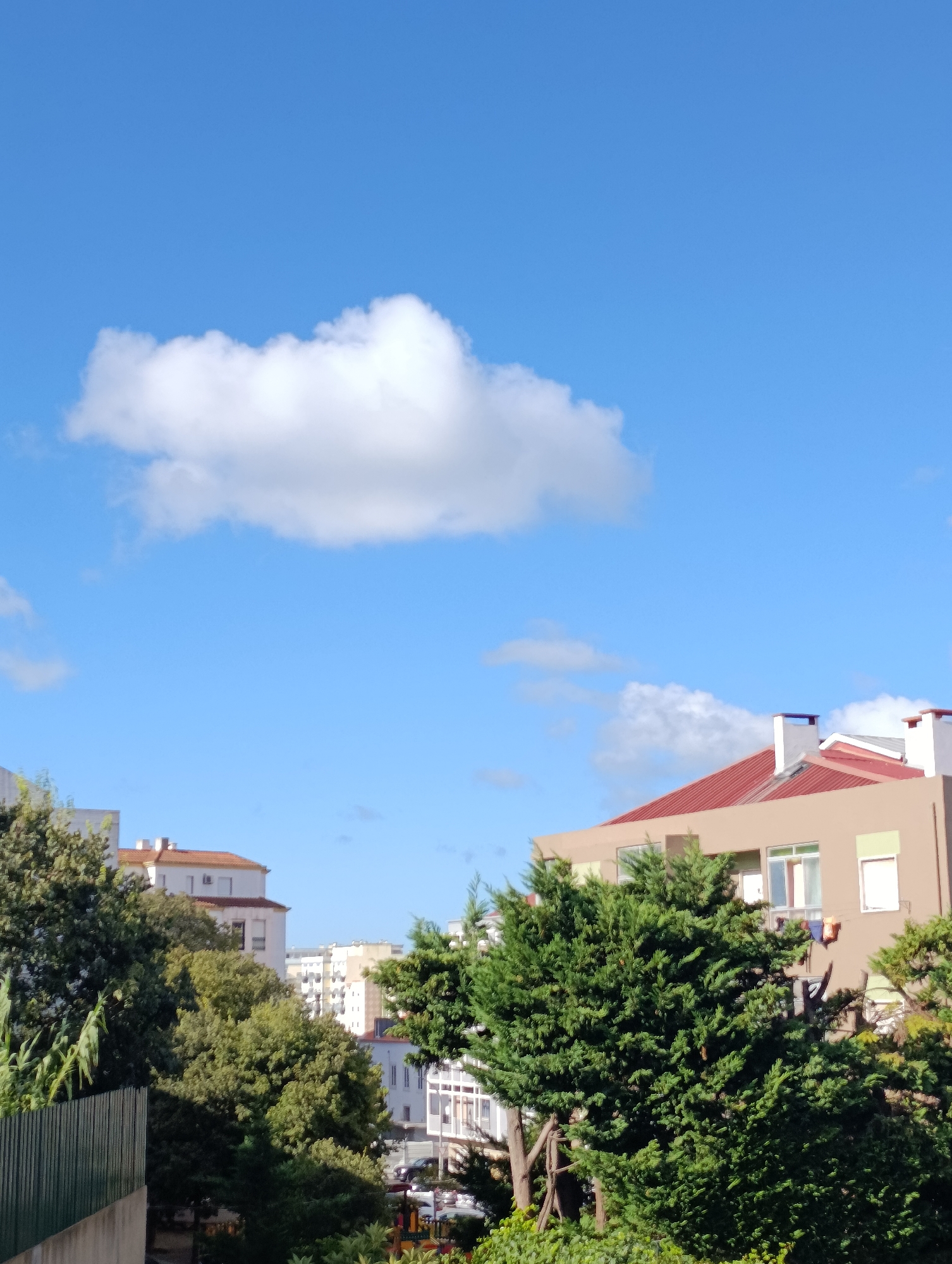 The image captures a clear blue sky with a prominent white cloud situated in the upper left quadrant. Below the sky, various buildings and lush green trees dominate the scene, suggesting an urban or suburban setting. In the foreground, a metal fence is partially visible on the left. Several apartment buildings with different architectural styles are discernible, including one with a reddish-brown roof and beige walls, featuring windows and chimneys. Another building in the background appears to be a taller, lighter-colored structure. Dense foliage from trees partially obscures some of the buildings. The presence of laundry hanging from a window on one of the buildings provides a domestic detail. The overall impression is of a bright, sunny day in a Portuguese city.