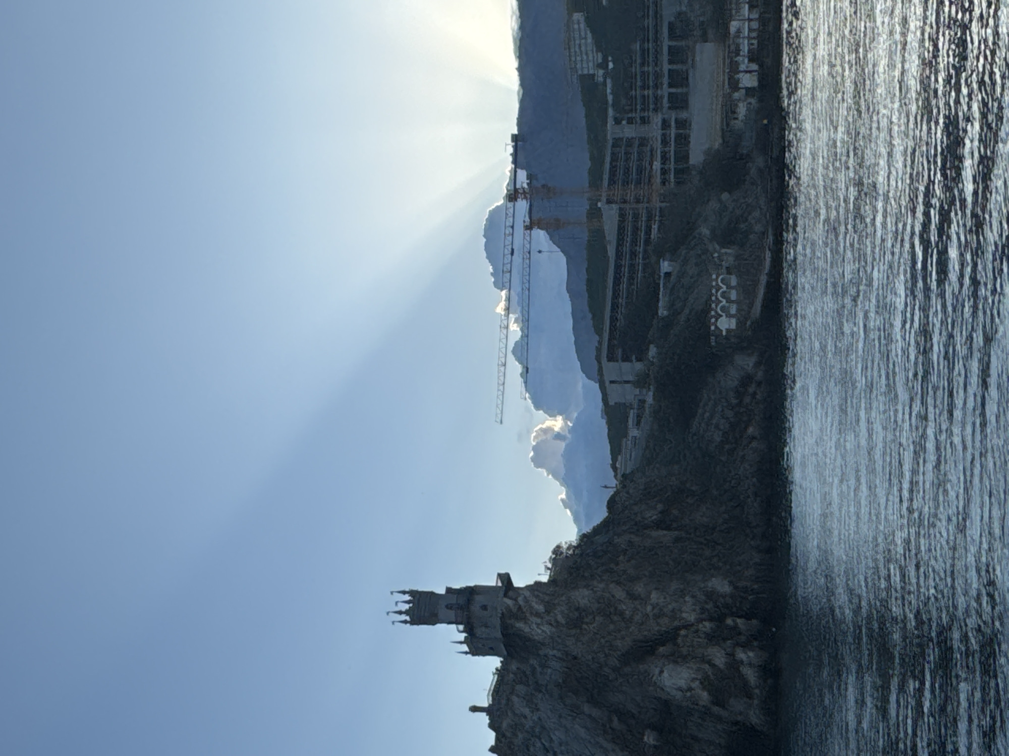 The image shows the Swallow's Nest castle perched on a cliff overlooking the sea, likely in the Гаспринский поселковый совет area of Ukraine. Construction cranes are visible in the background, suggesting development activity. The sky is clear with sun rays emanating from behind a cloud formation. The sea is calm with visible texture on the surface.