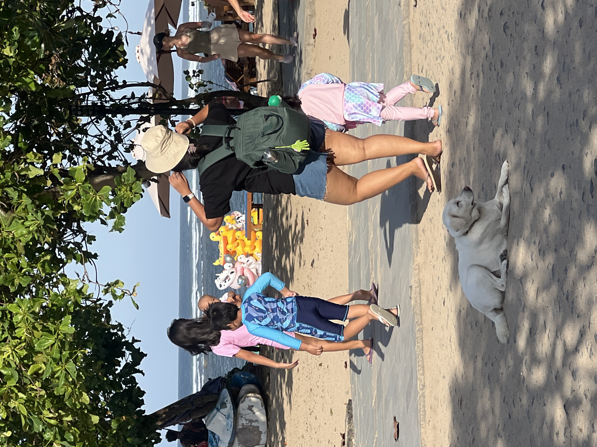 This media file depicts a scene at a beach in Denpasar, Indonesia. The sun is shining brightly, suggesting a clear and warm day, likely mid-morning or early afternoon. Several people are present, including adults and children, enjoying the beach environment.
In the foreground, a woman wearing a hat and a backpack stands on the sand. She is looking down, possibly at something in her hands or on the ground. Beside her, a young child in a pink shirt is visible. Further along the sandy path, another child is seen wearing a blue and navy swimsuit with a pattern. A white dog is lying down on the sand to the right of the path, appearing relaxed.
In the background, there are more people, including two women who appear to be walking or standing near beach umbrellas. The ocean is visible behind them, with gentle waves. The beach is a mix of sand and a paved walkway, with some trees providing shade on the left side of the frame. Several yellow inflatable ducks are piled up on the sand near the water's edge, possibly for sale or for children's play. The overall atmosphere seems casual and family-oriented, typical of a beach outing.