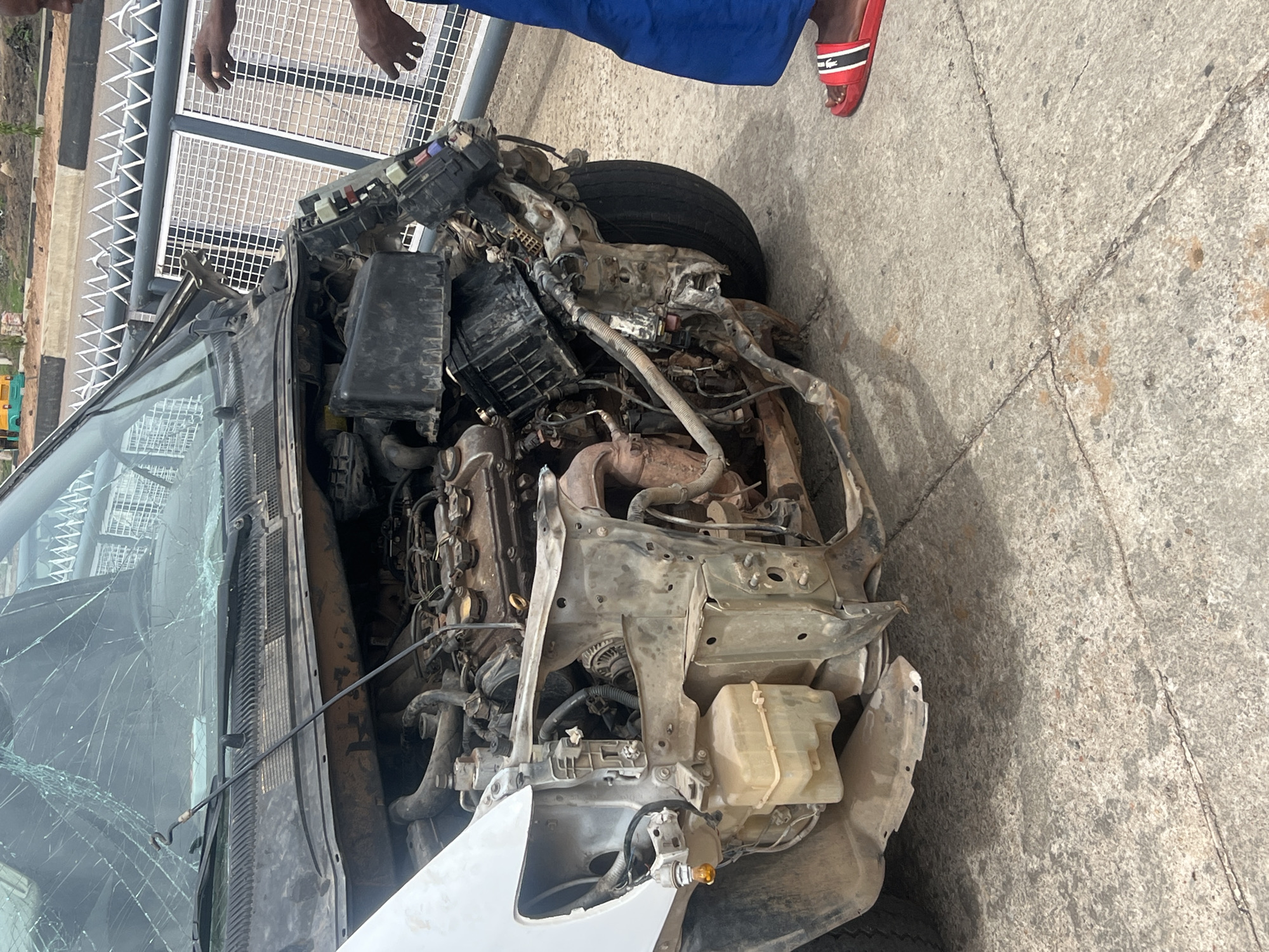 The image shows the front of a severely damaged vehicle, possibly a car, in Gura Top, Nigeria. The front of the vehicle is completely crushed, exposing the engine and internal components. The windshield is shattered. Two dark-skinned individuals are partially visible: one near the top-left corner with their hand visible, and another above the vehicle with a leg clad in blue and wearing a red sandal with a white logo, possibly Lacoste. The environment appears to be an open, paved area, potentially a roadside or parking lot. A metal railing is visible in the background.