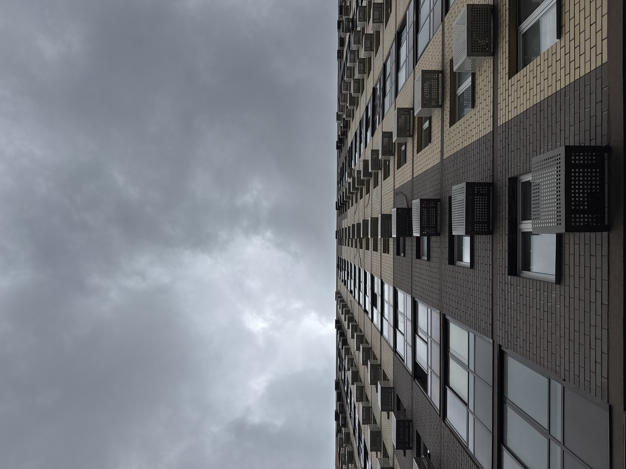 A low-angle shot captures the facade of a modern, multi-story apartment building under a dark, overcast sky. The building is constructed with alternating bands of light tan and dark brown brickwork. Numerous windows are visible, many reflecting the gloomy sky. Attached to the facade, at regular intervals, are many rectangular, mesh-covered units, likely air conditioning units. The perspective draws the eye upwards towards the vast expanse of clouds, creating a sense of scale and the imposing nature of the architecture. The scene appears to be in Мирный, Russia, based on the provided context.