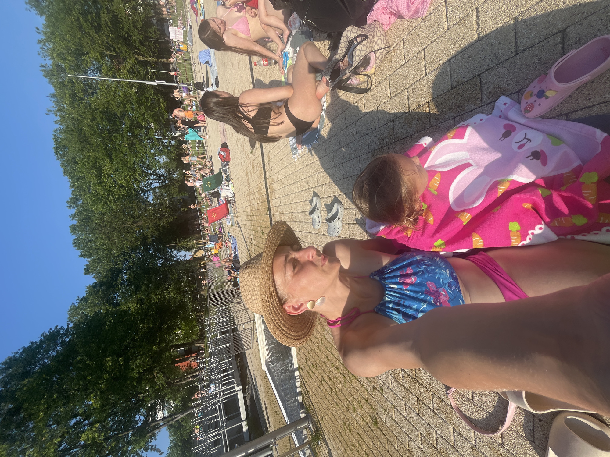 The image depicts a sunny day at an outdoor swimming pool or water park. In the foreground, a woman wearing a straw hat, a blue and pink floral bikini, and large earrings is lying down, looking up at the sky. Beside her, a young child is covered with a pink blanket adorned with a cartoon bunny and carrots. A pink clog and a pair of white Crocs are visible near them.
Further back, other people are sunbathing on lounge chairs and towels, and some are walking around. The ground is paved with light-colored bricks. Lush green trees line the background, and a clear blue sky is visible. A metal fence separates the paved area from what appears to be the pool or a grassy area. The lighting suggests it is daytime, likely midday, given the strong shadows. The overall atmosphere appears relaxed and recreational, indicating a leisurely day at a public facility. There is no visible text in the image.