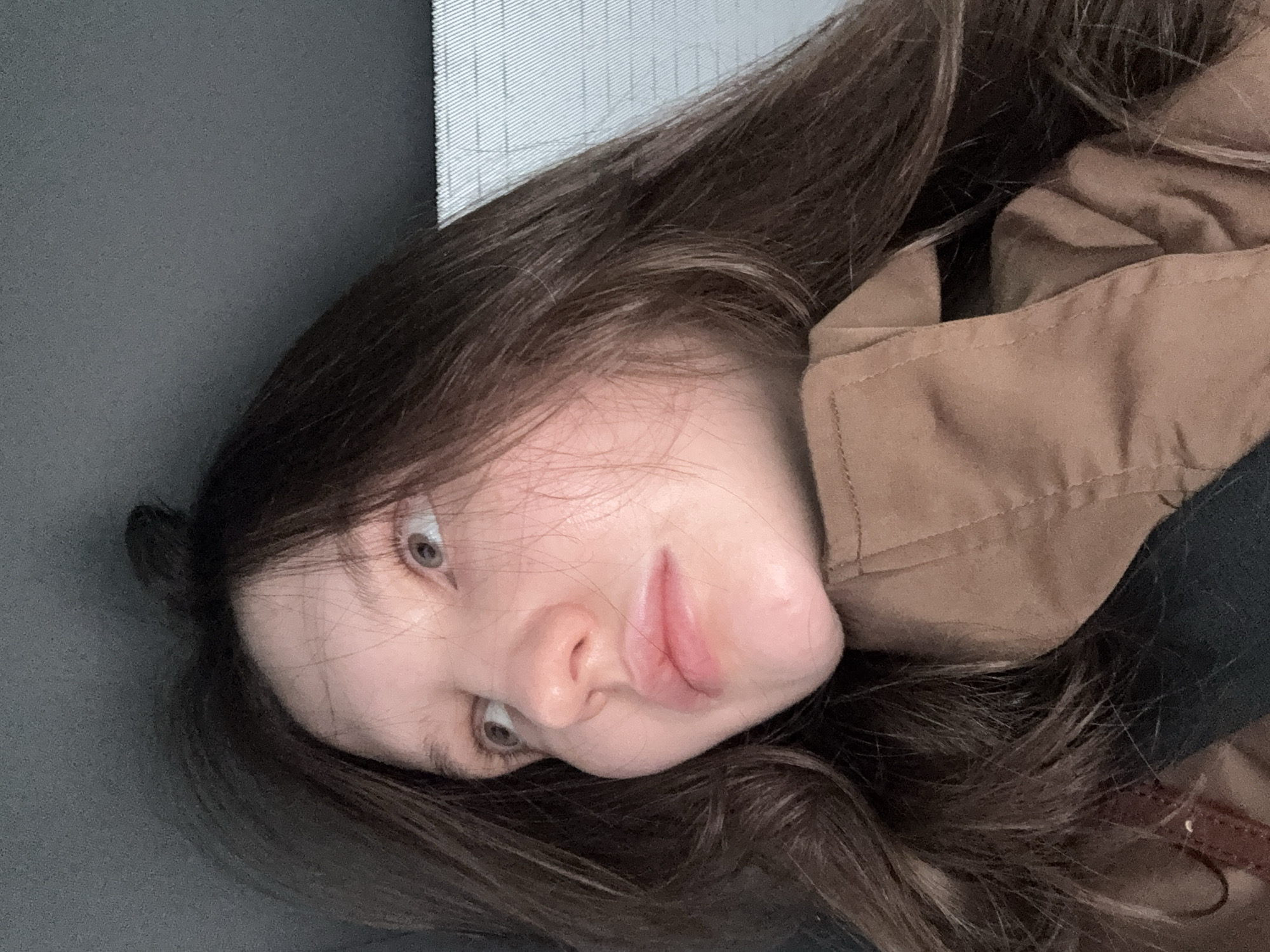 A young woman is positioned lying down, facing the camera with her head tilted towards the left. Her long, dark brown hair is loose and surrounds her face. She has fair skin, light-colored eyes, and slightly parted lips. She is wearing a tan-colored jacket with a dark collar. The background consists of a gray surface, possibly a seat or wall, and a textured gray and white area behind her head. The location cannot be ascertained from the image. There is no evidence the image was taken in Khimki, Russia.