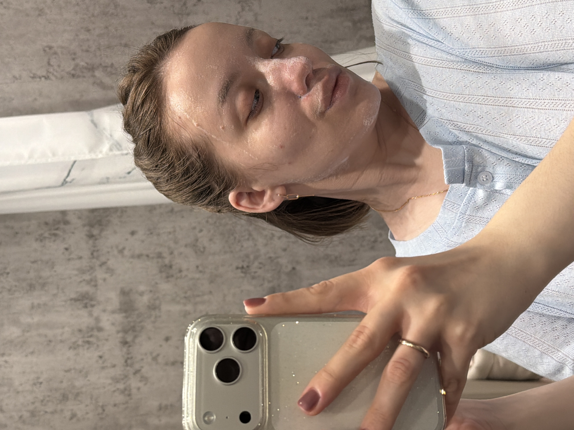 A woman is depicted taking a selfie indoors in Reshetnikovo, Russia. The image is rotated 90 degrees clockwise, showing her head tilted to the right. She has a white facial product, likely a mask or cleanser, applied to her face, prominent on her cheeks, nose, and forehead. Her medium-brown hair is pulled back, appearing sleek.
She wears a light blue, short-sleeved top with a textured, perforated pattern and buttons at the neckline. A delicate gold chain necklace is visible, along with a simple gold chain-link earring in her right ear. A gold band ring adorns her left ring finger. Her right hand, featuring dark pink nail polish, holds a silver/light-colored iPhone, which is protected by a clear, glittery case. The phone's triple camera lenses are clearly visible.
The background consists of a grey, textured wall, possibly concrete, and a white element, which might be a shower curtain with a faint marble pattern or a bathroom fixture. The lighting is bright and even. The woman's expression is neutral, with her eyes slightly closed or gazing downwards. No visible text is present in the image.