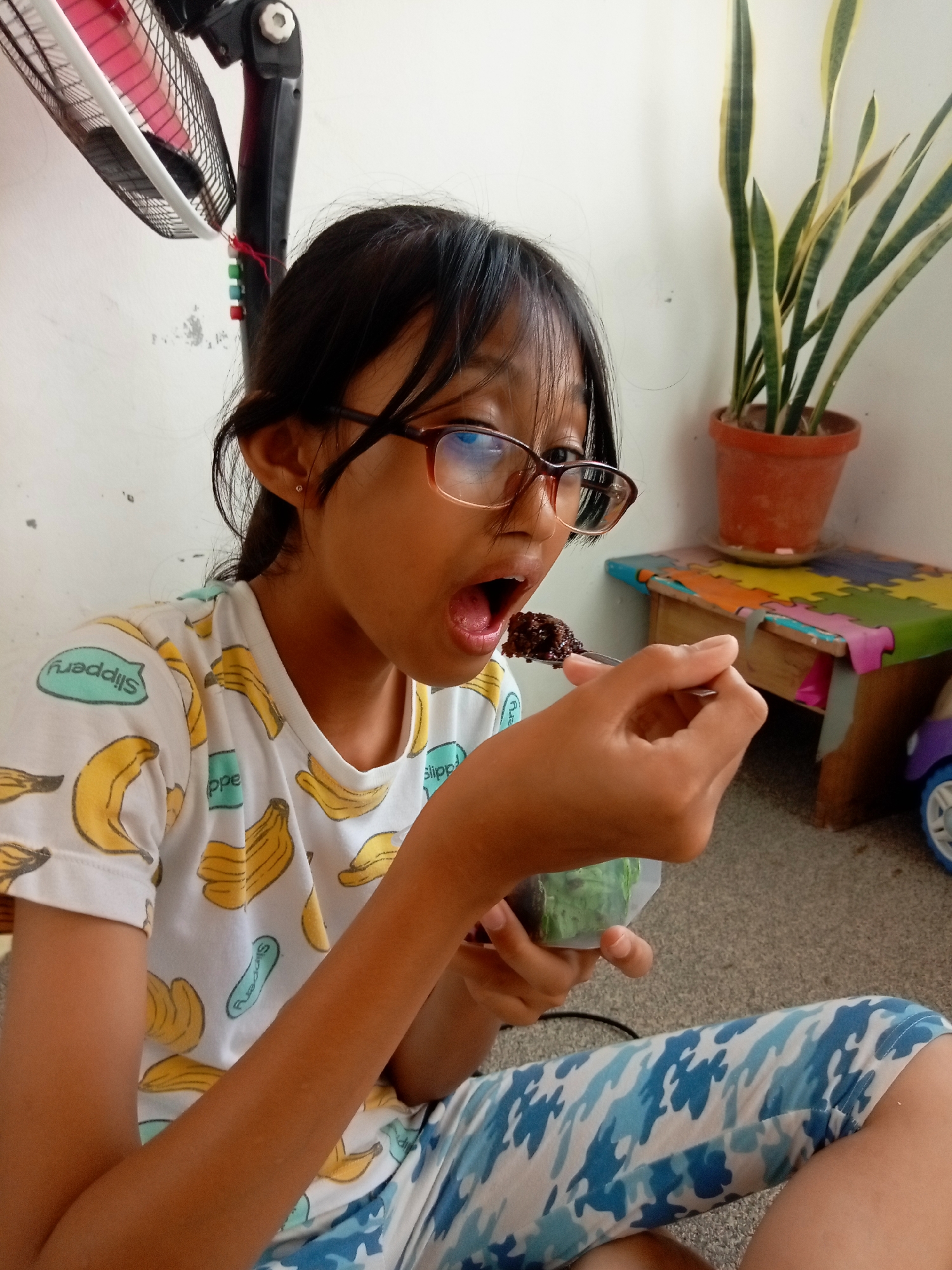 A young girl with glasses and dark hair is sitting on a carpeted floor in what appears to be a residential setting. She is wearing a white t-shirt with a banana print and blue camouflage-patterned pants. The girl is in the process of eating something dark brown from a spoon, with her mouth open. She is holding a small, clear container in her other hand, which appears to contain green material.
In the background, there is a tall, oscillating fan, a potted snake plant, and a low wooden table with a colorful, puzzle-piece-like covering. A small, toy-like object with a blue wheel is visible on the right side of the table. The wall behind the subjects is painted white and shows some minor scuff marks. The lighting suggests an indoor environment, possibly with natural light coming from an unseen source. The presence of a snake plant, oscillating fan, and simple furniture suggests a common domestic interior, not specifically indicative of a particular district within Batam, Indonesia.