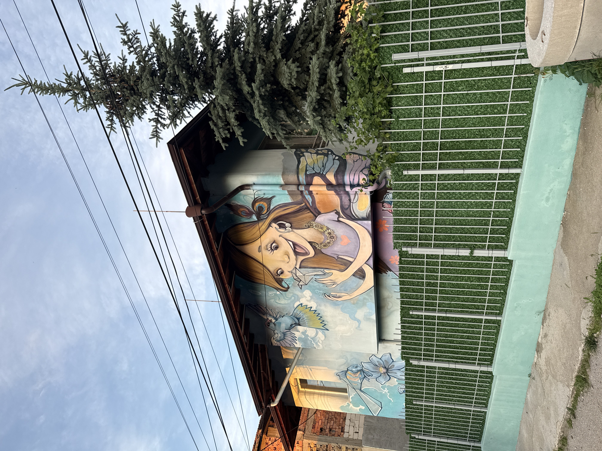 In an urban setting in an unknown city, a mural is painted on the side of a building. The mural depicts a cartoonish girl with blonde hair and large eyes, holding a small origami crane. She has butterfly wings and is surrounded by birds and flowers. The building is a two-story structure with a brown roof and a light blue wall. A tall evergreen tree stands to the right of the building, partially obscuring it. A green fence, topped with artificial turf, runs along the front of the property, separating it from a concrete sidewalk and a dirt path. Overhead, several power lines stretch across the sky, which is partly cloudy. The scene appears to be captured at dusk, with the soft lighting casting a warm glow on the mural.