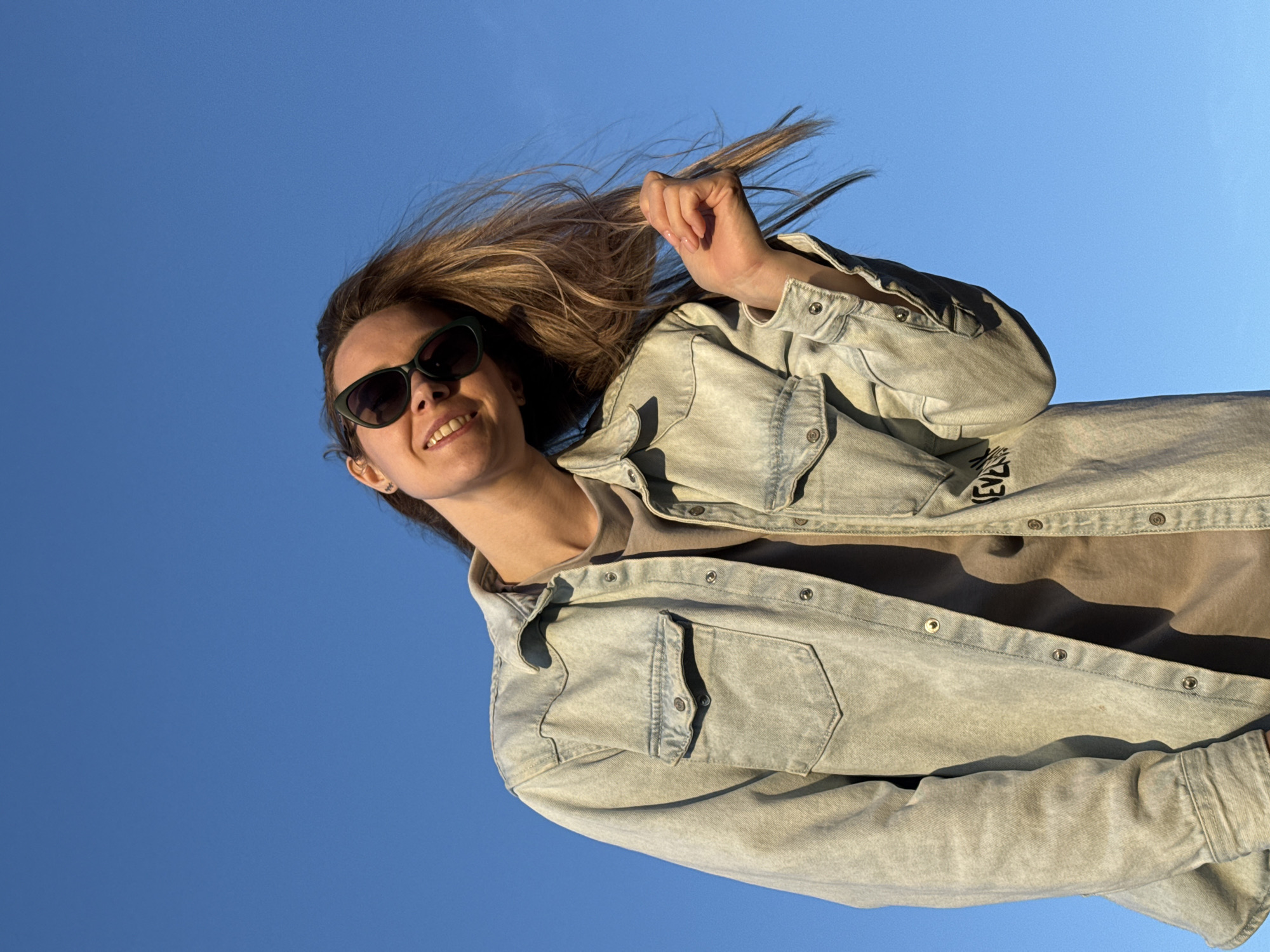 A woman is outdoors, seen from a low angle with a clear blue sky as the background. She is smiling and wearing sunglasses and a light-colored denim jacket over a neutral-colored top. She is holding a strand of her light brown hair in her right hand. The image was taken in Chaikovsky, Russia.