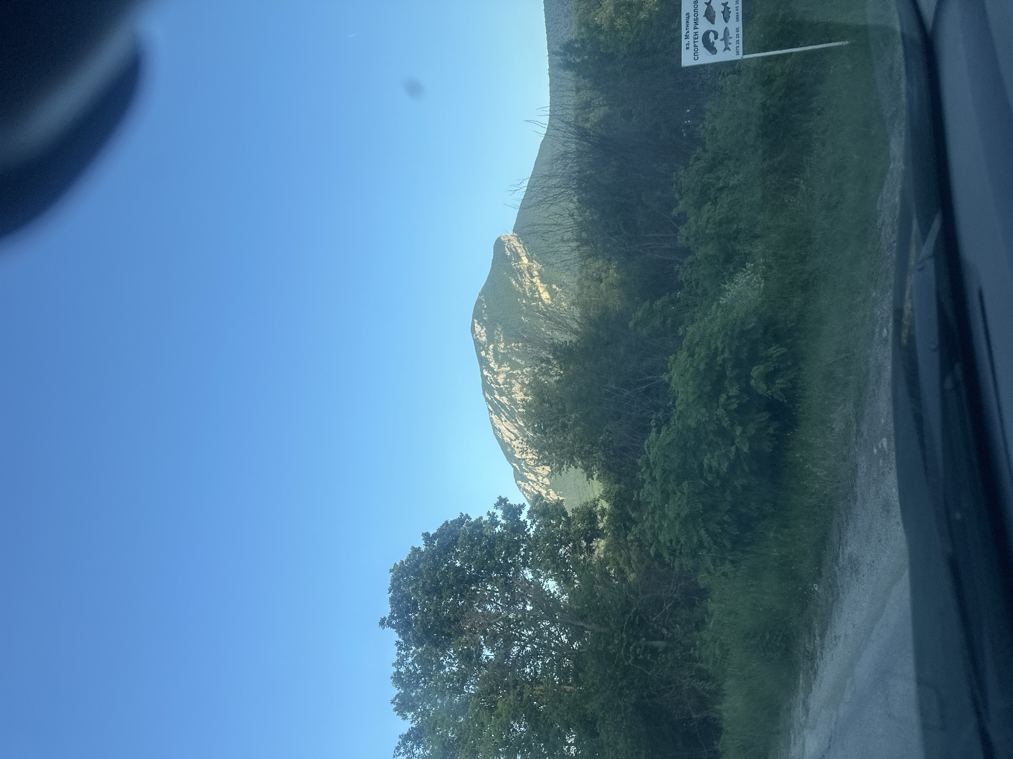 The media file captures a serene mountainous landscape under a bright, clear blue sky, seen from inside a vehicle. The primary subject is a large, rugged mountain or hill dominating the middle ground, its slopes covered in dense green vegetation with visible rocky, lighter-colored sections near its peak.
In the foreground, to the left, is a vibrant green tree with full foliage, and across the bottom and right side, more lush green bushes and trees create a natural boundary. A light-colored dirt or gravel road is visible along the right edge, hinting at a route through this natural setting. Part of a vehicle's interior (likely a window frame or pillar) is visible on the far right, suggesting the image was taken from a car or similar transport. A small, blurry, dark shape is present in the upper left corner, possibly a reflection or another part of the vehicle's interior. A faint white streak, possibly a distant airplane contrail, can be seen in the sky towards the upper right.
No people are visible in the image. The scene suggests an activity of travel or observation of nature, likely while driving through or near Glavatsi, Bulgaria.
Notable details include the bright daylight, indicating clear and sunny weather, likely during the morning or afternoon. The overall atmosphere is peaceful and natural.
A white sign is partially visible in the upper right quadrant. The Cyrillic text on the sign reads:
- "ВЗ. Мътница" (VZ. Mutnitsa, potentially a place name or area)
- "СПОРТЕН РИБОЛОВ" (Sports Fishing)
Below this text, there are graphic icons of various fish. Two phone numbers are also visible at the bottom of the sign: "0879 23 20 03" and "0884 83 38".