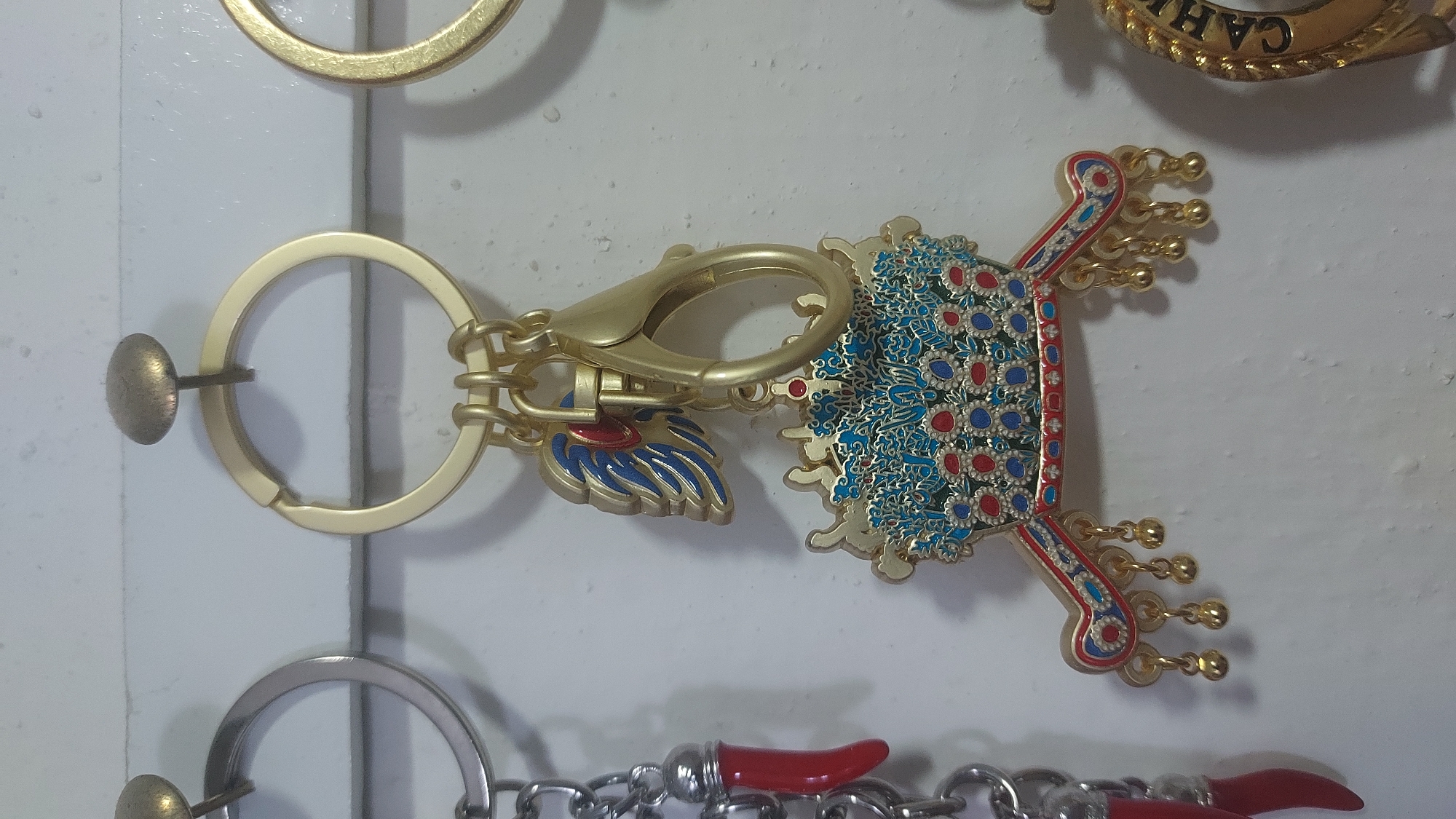 The image displays a close-up view of several decorative keychains hanging on a white wall, possibly in a market or shop. The central keychain features an ornate crown-like ornament with intricate blue and red enamel detailing, gold trim, and small dangling gold balls. It is attached to a gold keyring via a clasp and a small blue and red winged charm. To the left of the central keychain, another keychain is partially visible, featuring a silver ring and a dangling red chili pepper charm on a silver chain. To the right, a portion of a gold keychain with embossed lettering is visible. The keychains are suspended by nails or hooks. The lighting is somewhat harsh, casting shadows and highlighting the textures of the wall and the metallic surfaces of the keychains. No people or animals are present in the frame, and no specific elements suggest the location is Kogon City, Uzbekistan.