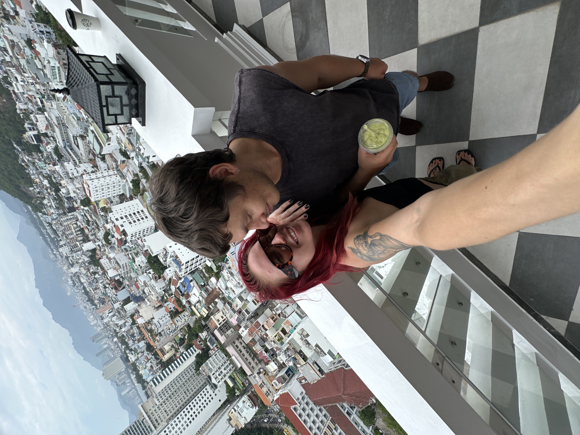 A couple, a woman with bright red hair and sunglasses, and a man with brown hair, are taking a selfie on a balcony overlooking a dense, urban cityscape in Nha Trang, Vietnam. The woman is leaning into the man, who is holding a green drink. The balcony floor is tiled with a black and white checkered pattern. In the background, mountains can be seen in the distance, shrouded in haze. The architecture of the city includes a mix of modern high-rises and older, lower-rise buildings with red-tiled roofs. A traditional-style lantern sits on the white railing of the balcony. The sky is overcast.