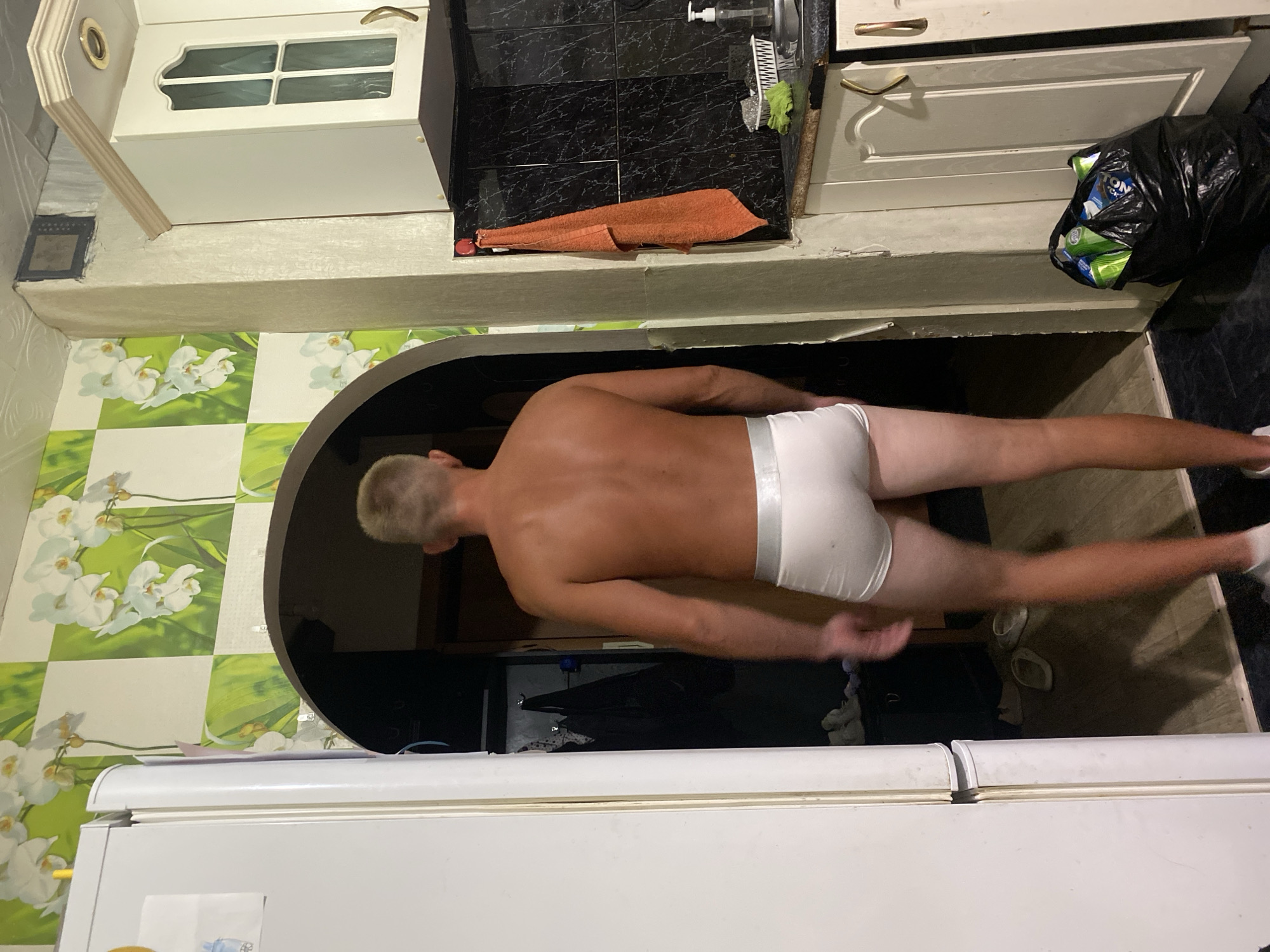 A shirtless man wearing white underwear is reaching through an arched doorway in a kitchen. He has short blond hair and a tanned back. The kitchen has green and white floral tile on the walls. A white refrigerator is in the foreground. Above the doorway are white cabinets, some with glass panes, and a black tiled backsplash. A black trash bag full of cans is near the cabinets. An orange towel is draped on the countertop. The location is likely a residential kitchen; there is no information to identify that this image was taken in the city of Chaikovsky, Russia.