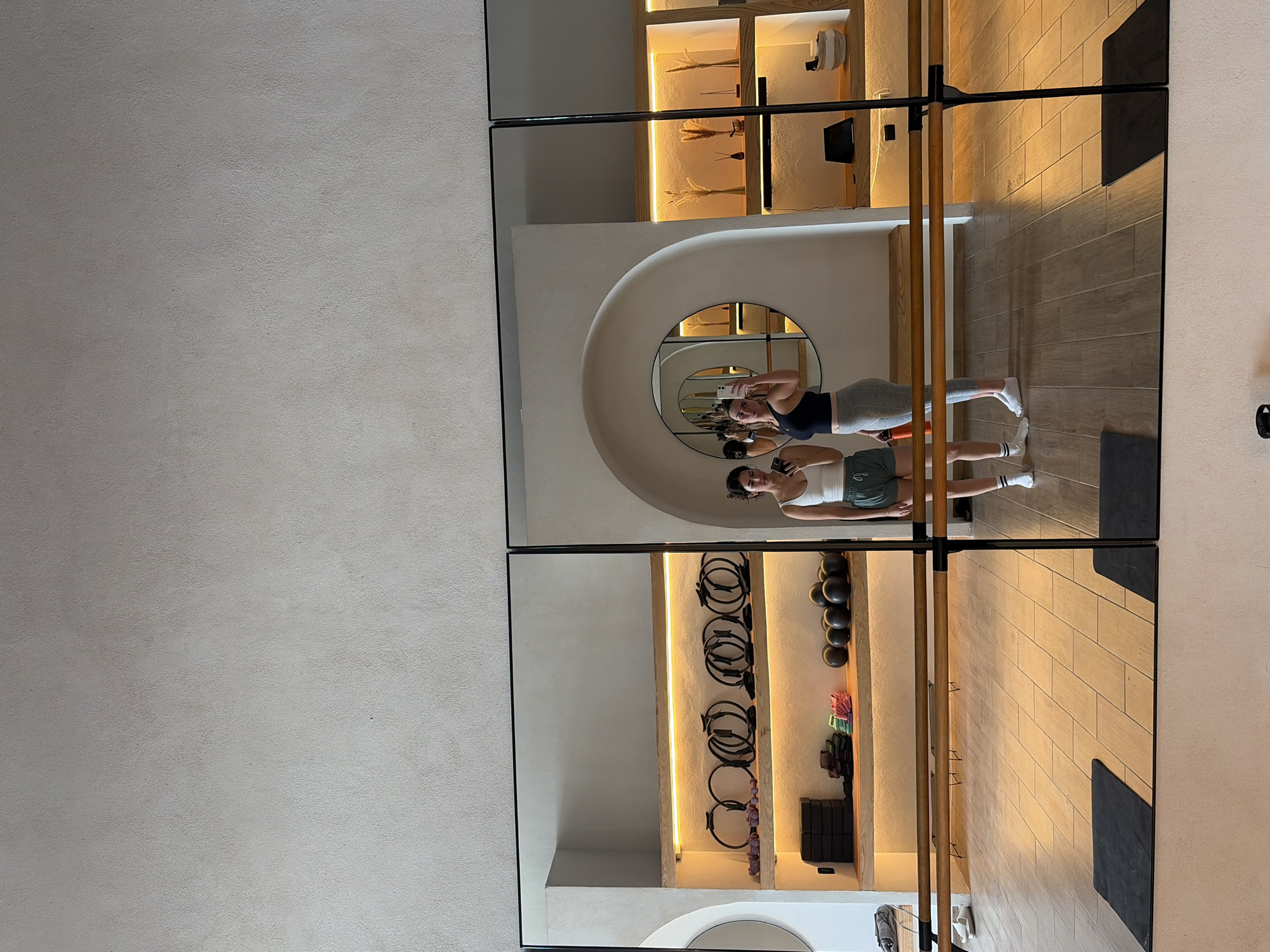 Scene in San Bernardino Tlaxcalancingo, Mexico, featuring a mirrored wall showing reflections of two women and portions of a fitness studio. The women are reflected in a large mirror panel that is divided into quadrants, with each section reflecting parts of the studio and the individuals. One woman, slightly taller, is taking a selfie in the mirror, wearing a dark top and light-colored leggings. The second woman, wearing a light top and dark shorts, is standing next to her. The studio features shelves with various workout equipment, including resistance bands, weights, and yoga mats. The flooring is composed of light-colored rectangular tiles, and there is a ballet barre running horizontally across the mirror. The architecture of the studio includes rounded alcoves and arches, giving the space a modern aesthetic.