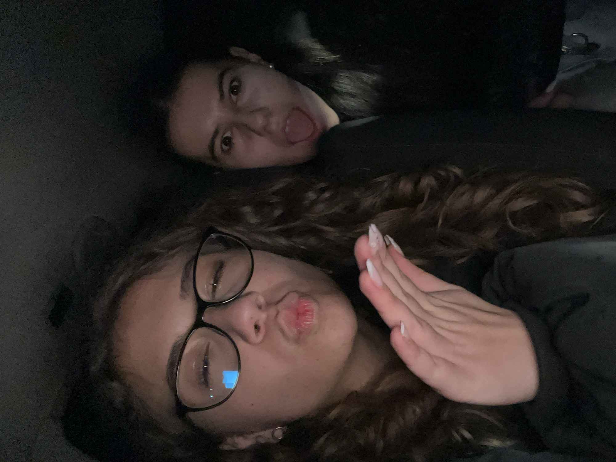 Two women are visible in the image. One woman, positioned in the foreground, has curly brown hair, glasses, and is pouting her lips. Her hand is raised to the side of her face, with long, manicured nails visible. The other woman, positioned in the background, has dark hair and is sticking her tongue out. The environment appears to be dark, possibly indoors. The location of Karavelovo, Bulgaria, is not evident in the image.