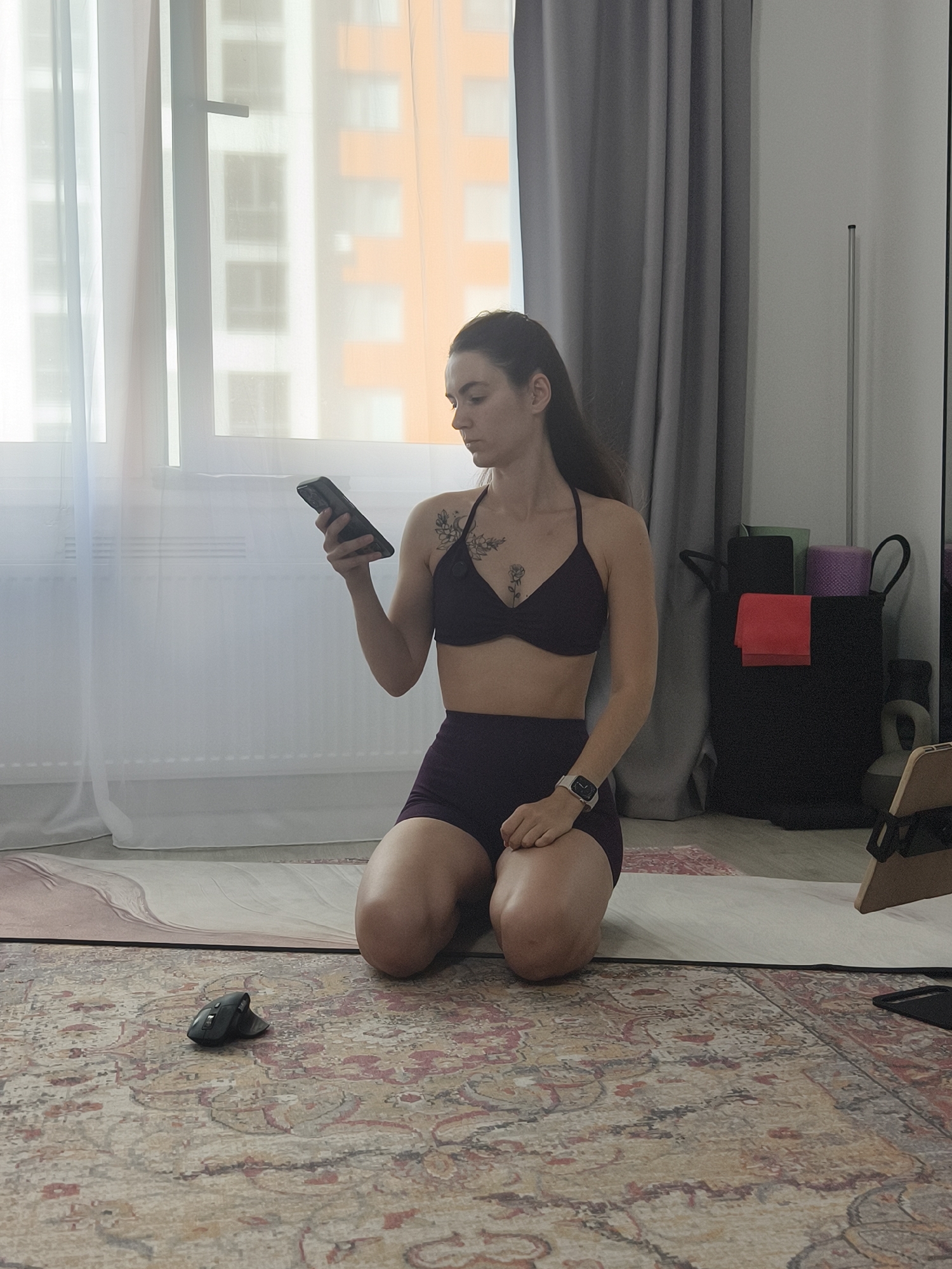 A full-shot, indoor, waist-up image shows a woman with long dark hair, wearing a dark purple sports bra and matching shorts, kneeling on a yoga mat. She holds a smartphone in her left hand and looks down at it. A tattoo of a rose is visible on her left shoulder blade. She wears a white Apple Watch on her left wrist. To her left, on a patterned rug, is a black computer mouse. Behind her, a window with sheer white curtains reveals a residential building. To her right, a grey curtain hangs, and a black hamper filled with colorful yoga mats and a red towel stands nearby. A light-colored yoga mat is visible behind the woman, and a wooden stand with a tablet is on the floor to the right. The scene appears to be a home setting, likely a living room or dedicated exercise space, and there is no indication of the specific city of Tyumen, Russia.