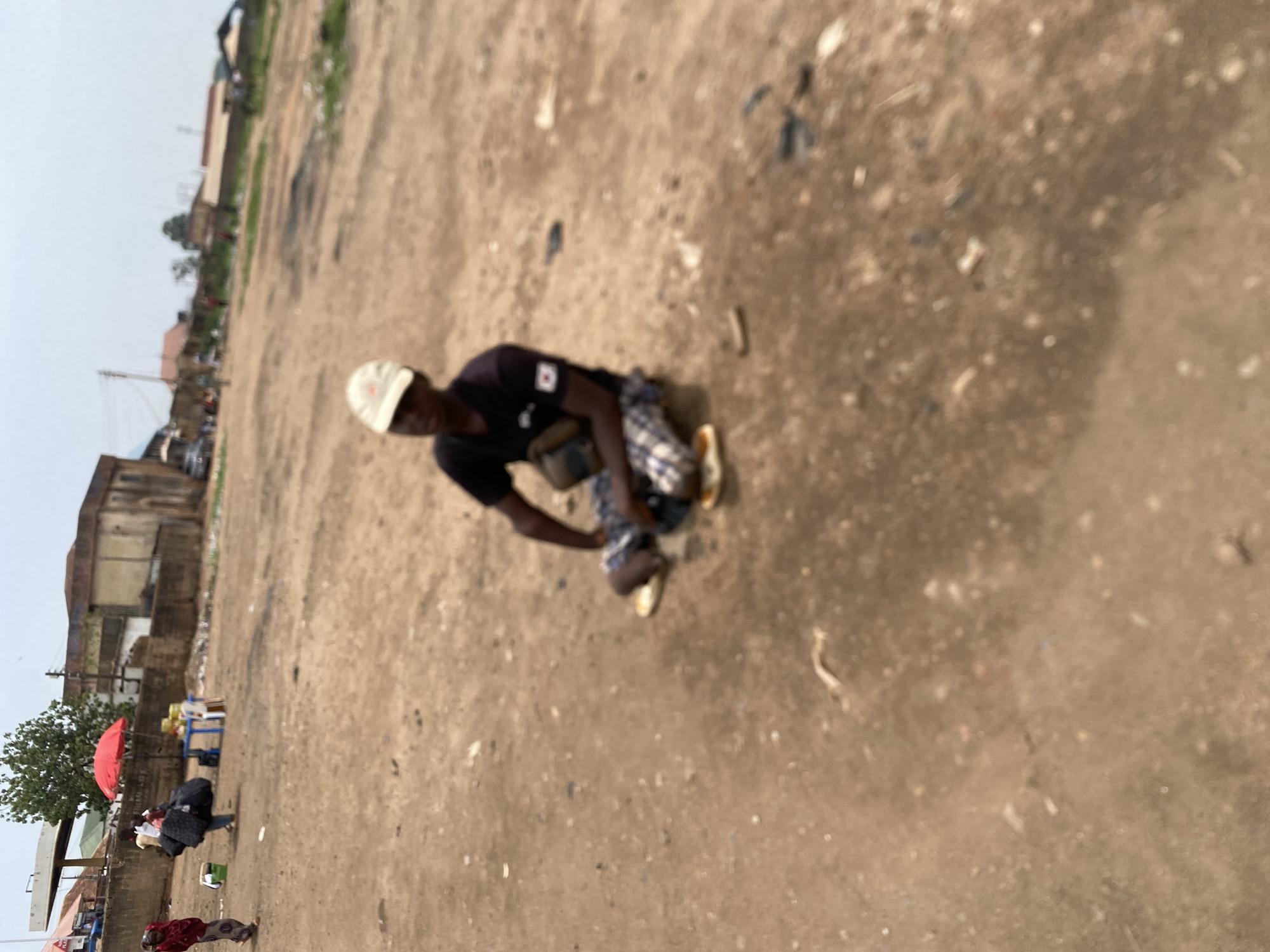 A blurry, ground-level, medium shot depicts a man crouching outdoors on a dirt and gravel surface in Bukuru, Nigeria. The man, who is African, is wearing a dark t-shirt, plaid pants, and light-colored shoes and a light-colored cap. He appears to be eating food from a plate or container. The background includes indistinct buildings and trees and other people in the distance. The sky is overcast. A vendor with a red umbrella is visible in the distance.
